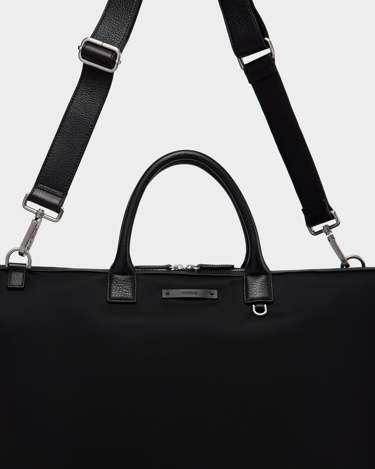The Rudsak DUBLIN - 9324514 Black is a canvas leather weekender tote with silver hardware, two short handles, and an adjustable, detachable shoulder strap displayed against a plain light background.