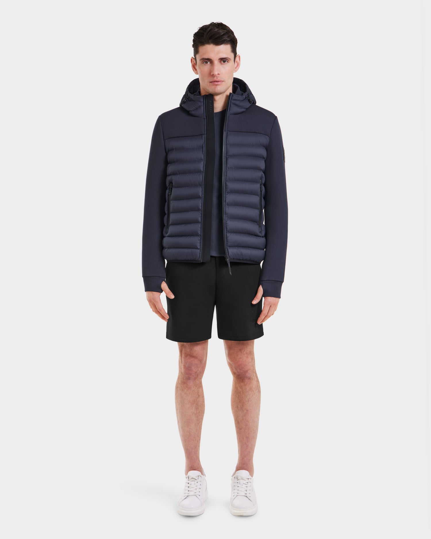 A man faces forward, wearing the Rudsak DURIN M - M123515 Navy mens hooded jacket with lightweight down insulation, paired with black shorts and white sneakers against a plain white background.