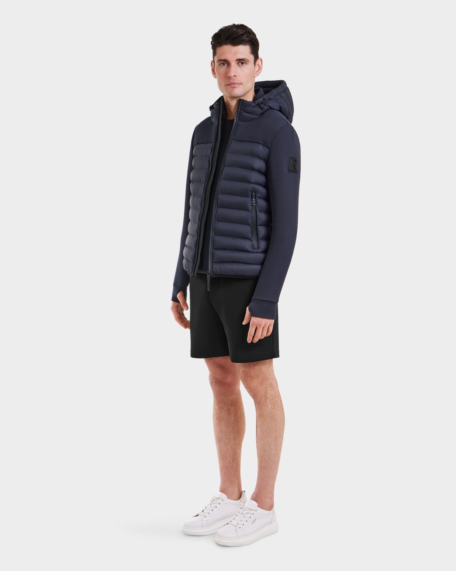 A man faces forward against a plain white background, wearing the Rudsak DURIN M - M123515 Navy mens hooded jacket with lightweight down insulation, paired with black shorts and white sneakers.
