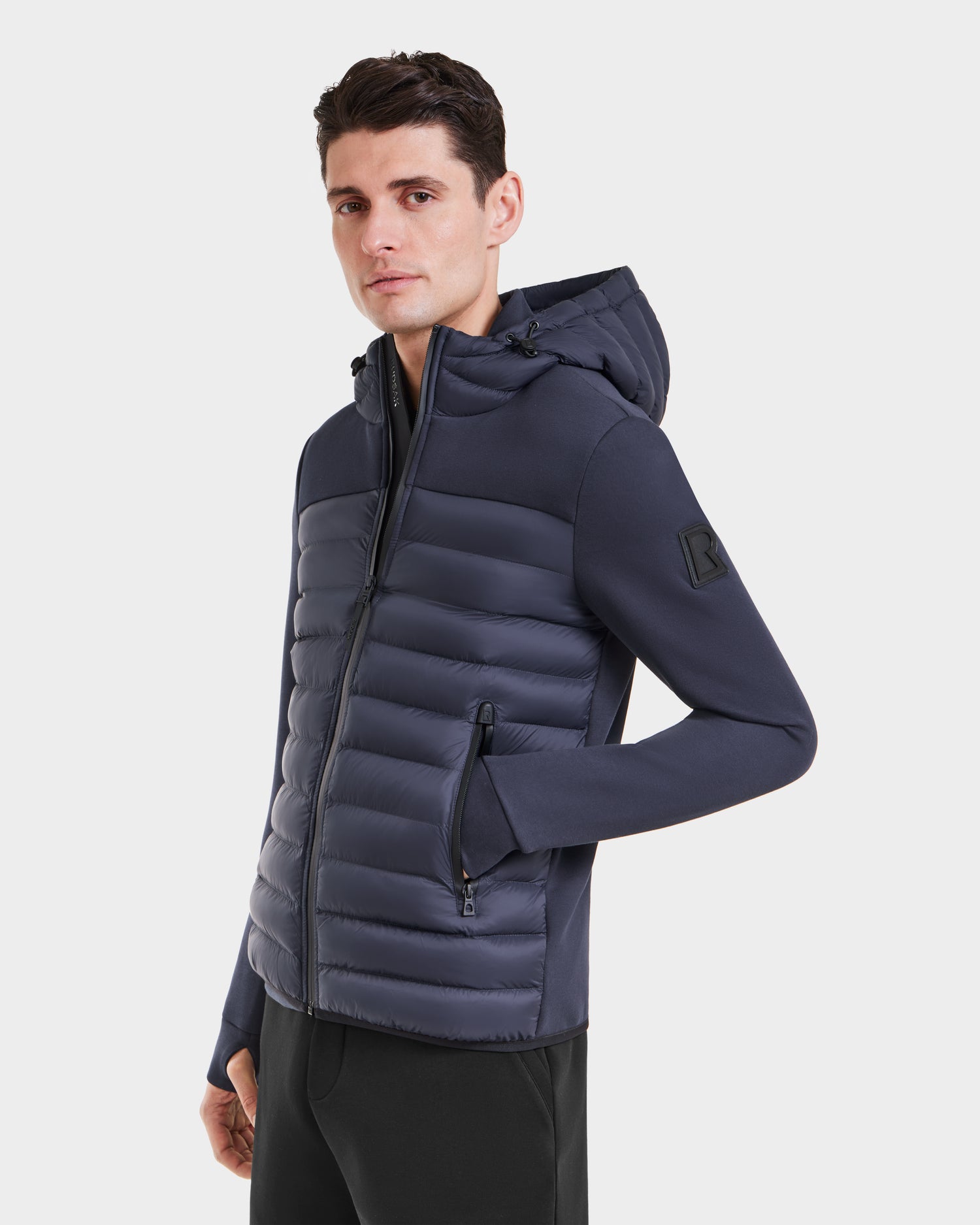 A man wears the Rudsak DURIN M - M123515 Navy, a fitted men’s hooded jacket with zippered pockets and lightweight down insulation, standing against a plain white background and looking slightly to the side.
