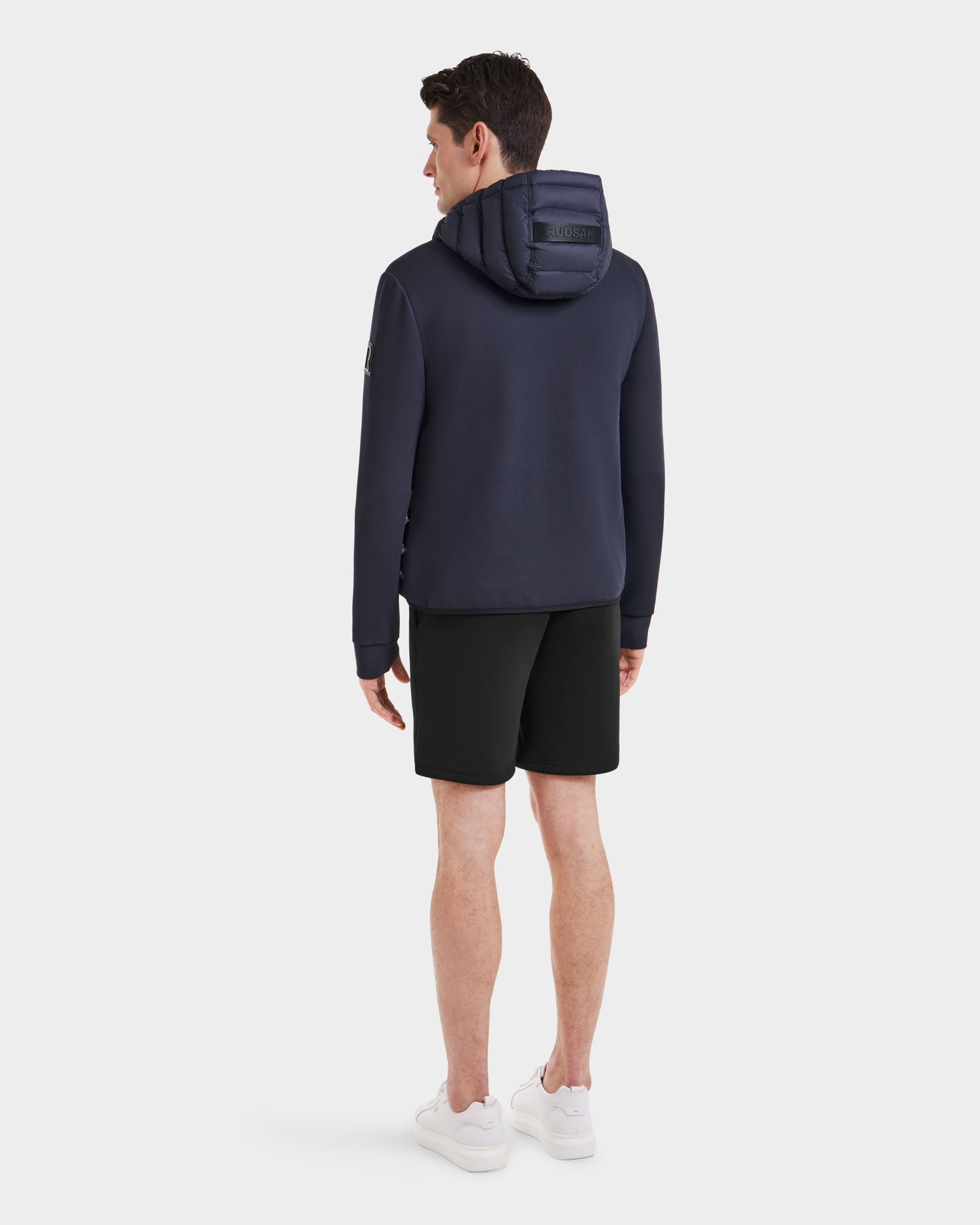 A man faces away, wearing the Rudsak DURIN M - M123515 Navy mens hooded jacket with lightweight down insulation, black shorts, and white sneakers against a plain white background.