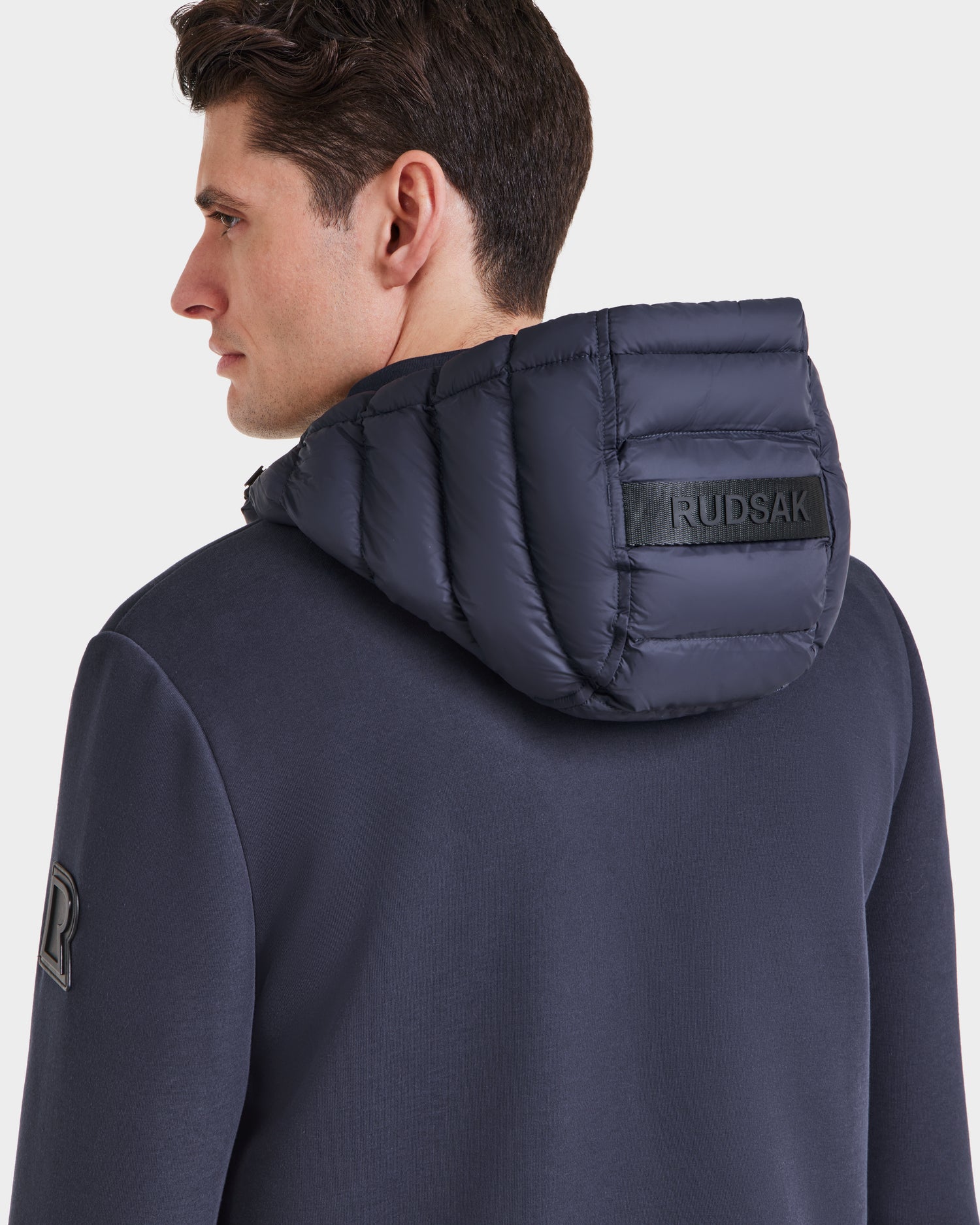 A man in a navy Rudsak DURIN M - M123515 men’s hooded jacket with lightweight down insulation and a quilted, padded hood featuring the Rudsak label faces away against a plain white background.