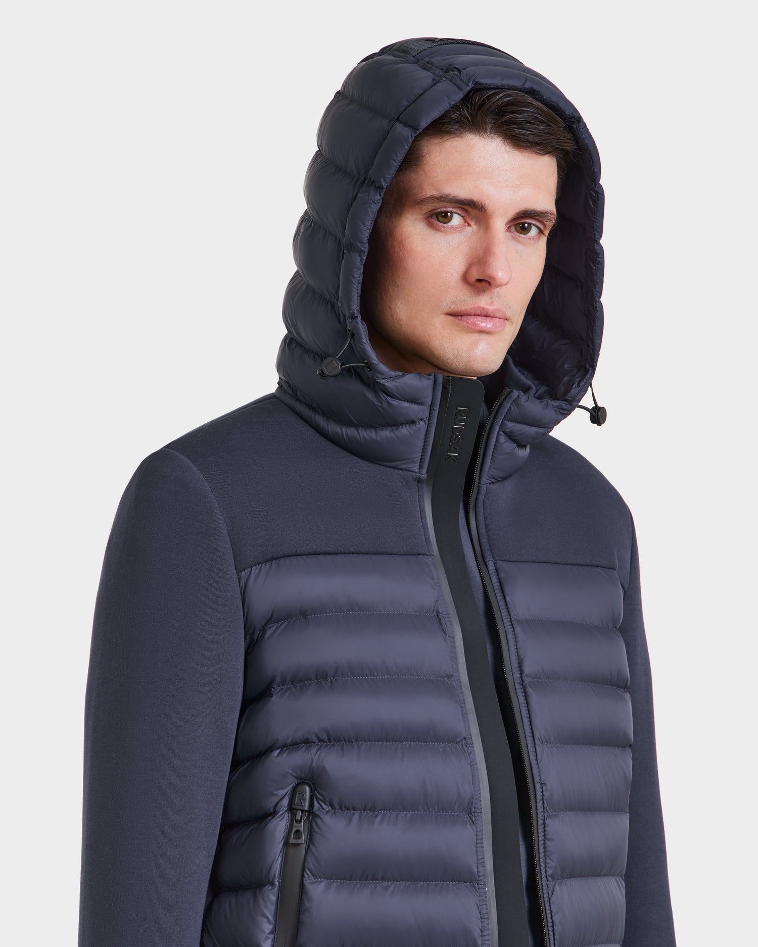 A man wearing the Rudsak DURIN M - M123515 navy mens hooded jacket with lightweight down insulation and zippered pockets, looking at the camera against a plain white background.
