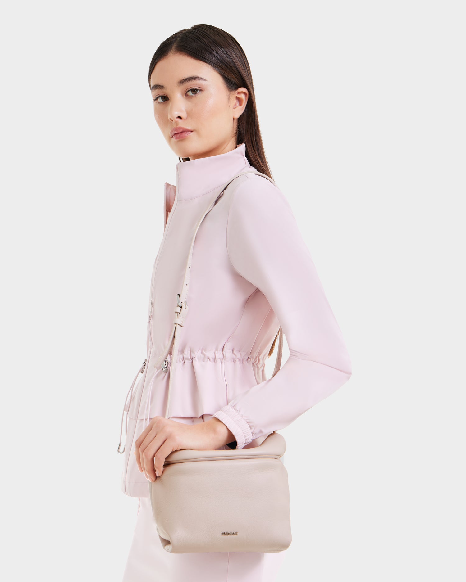 A woman with straight dark hair, wearing a light pink jacket, carries the Rudsak FANIA - 8325011 Soft Pink womens crossbody bag in light beige pebble leather. The minimalist bag stands out as she poses against a plain white background.