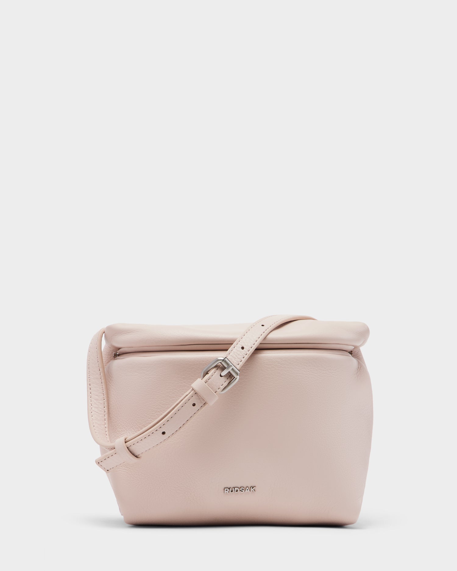 The FANIA - 8325011 Soft Pink by Rudsak is a womens crossbody bag in soft pink pebble leather, featuring a flap closure, adjustable strap, and subtle Rudsak branding on the front for a sleek minimalist look.