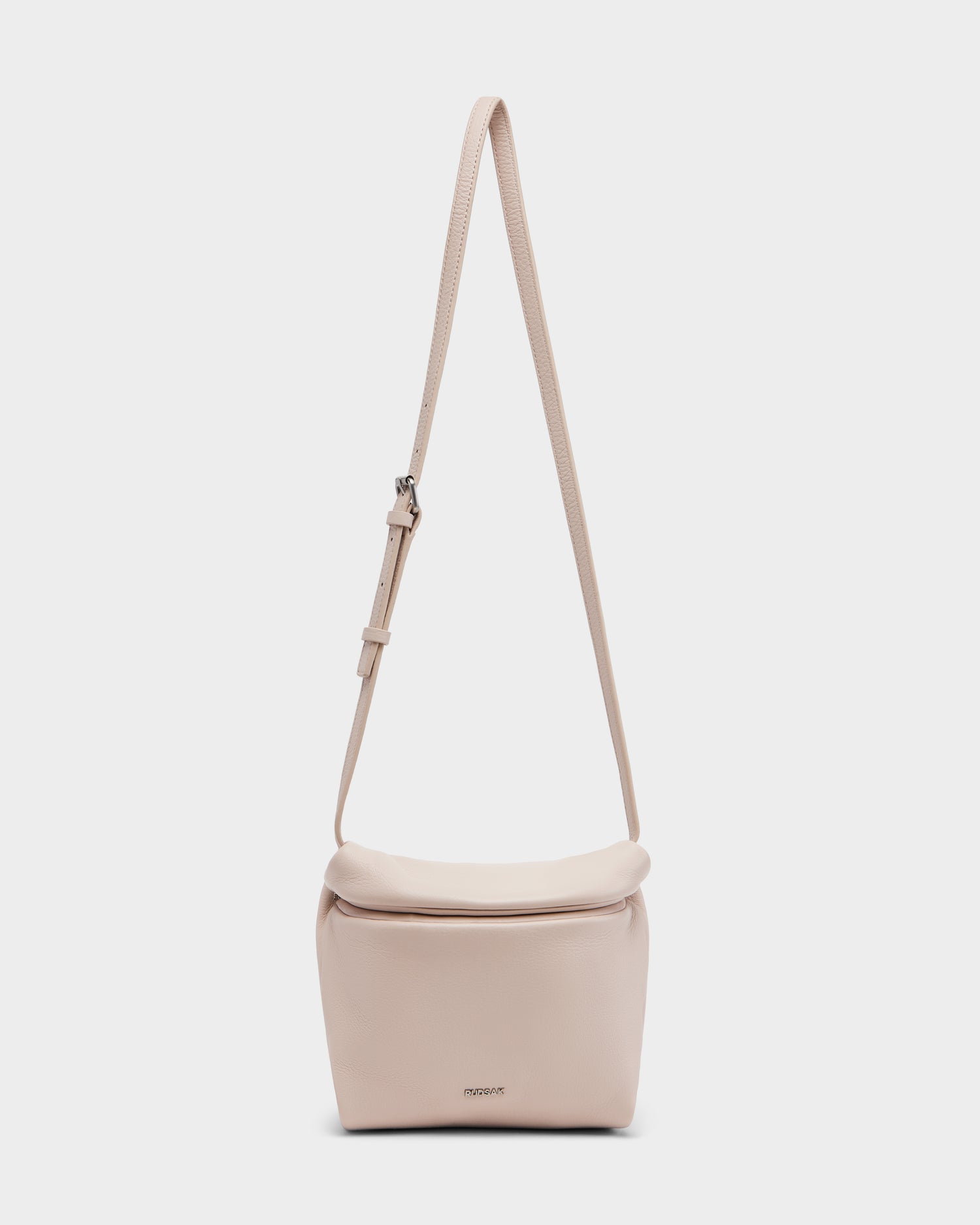 The Rudsak FANIA - 8325011 Soft Pink women’s crossbody bag features a minimalist design, adjustable strap, soft rectangular shape, small front logo, and is crafted from premium pebble leather against a white background.