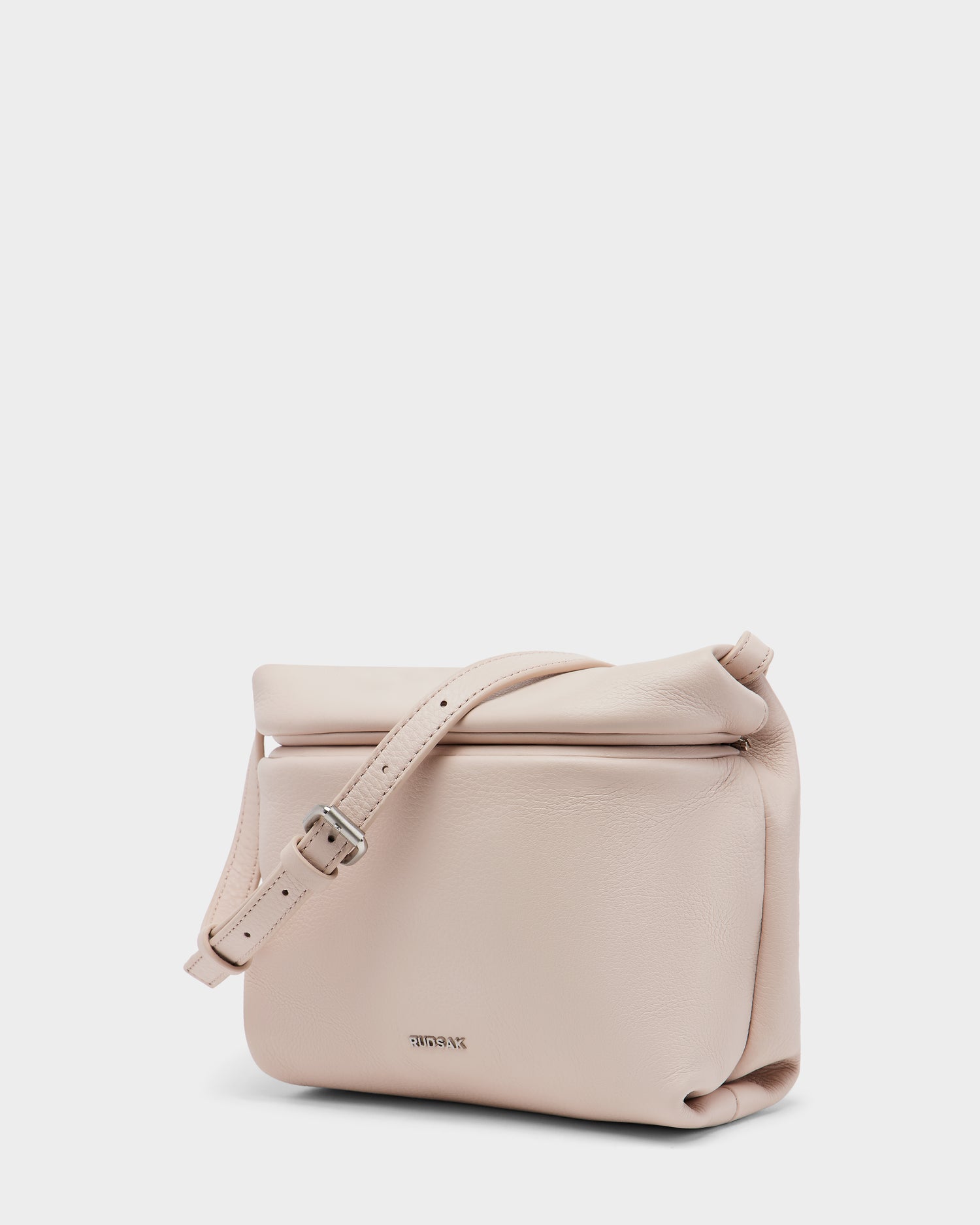 The FANIA - 8325011 by Rudsak is a soft pink womens crossbody bag in pebble leather, showcasing a minimalist design with a buckle-adjustable strap, curved edges, and a subtle metallic logo on the front.