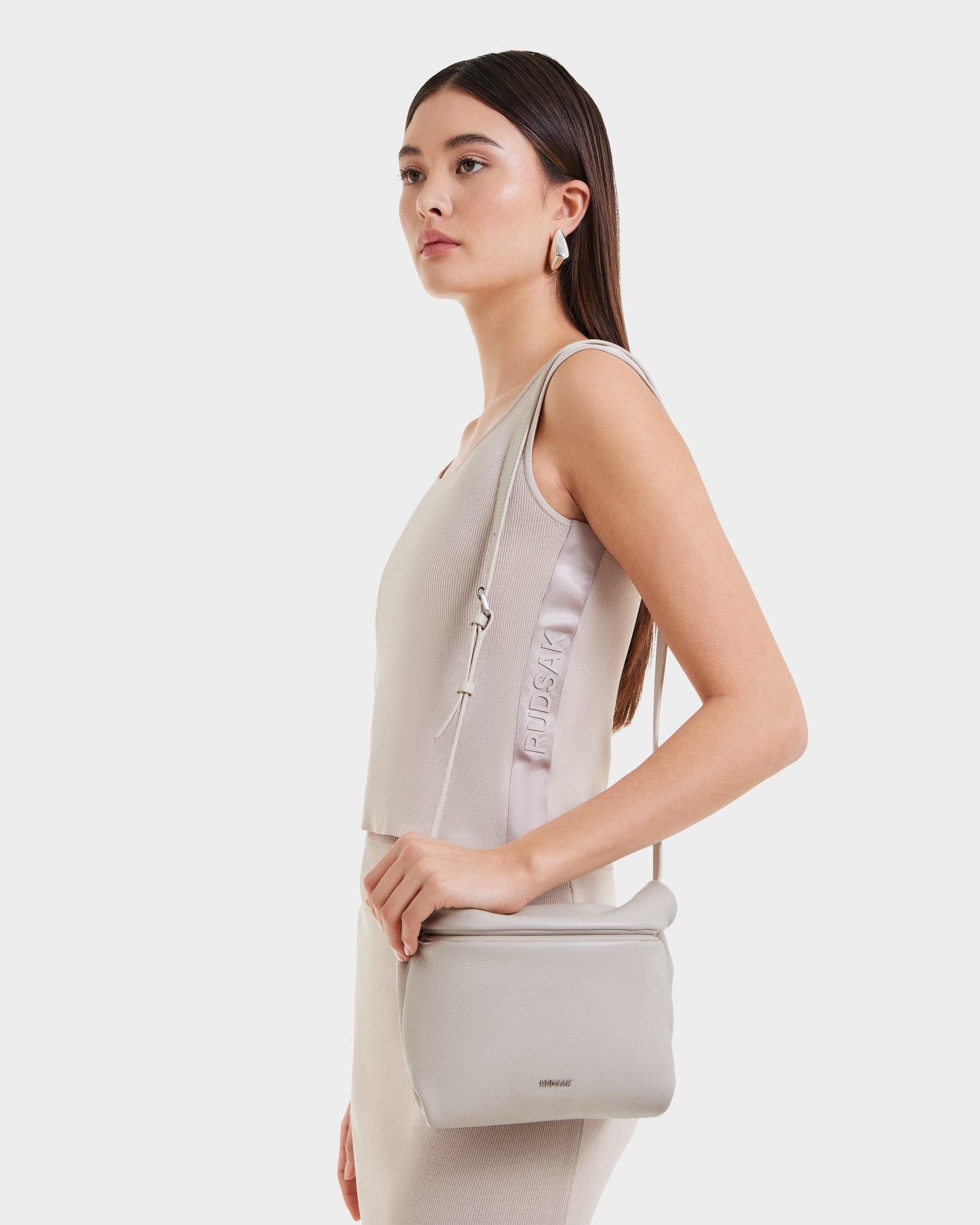A woman with straight brown hair wears a sleeveless beige outfit, holding the Rudsak FANIA - 8325011 Vanilla crossbody bag. Featuring a minimalist design, she stands against a plain white background, gazing slightly to the side.