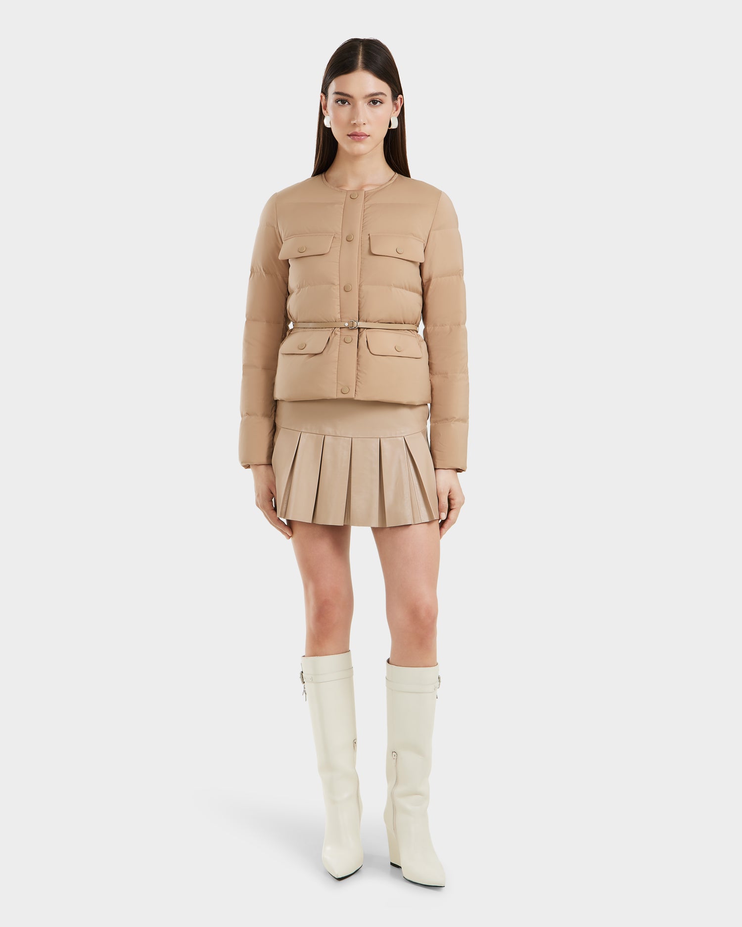 A woman faces forward in Rudsak’s FRANCESCA - 8125021 Almond beige quilted puffer jacket with a vegan leather belt, paired with a matching pleated mini skirt and knee-high white boots, set against a plain white backdrop.