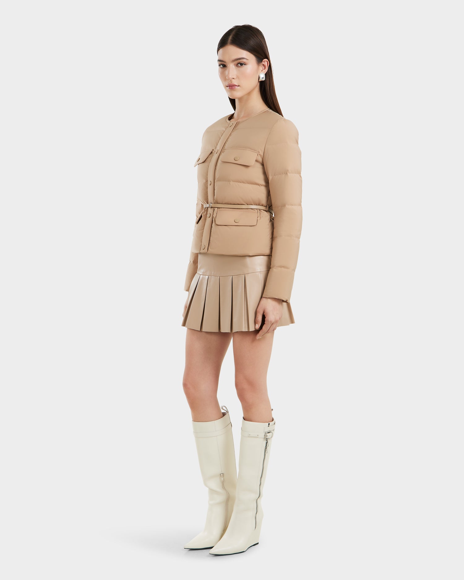 A woman stands slightly to the side against a plain white background, wearing Rudsak’s FRANCESCA - 8125021 Almond tan quilted puffer jacket with a vegan leather belt, a matching pleated skirt, and knee-high white boots.