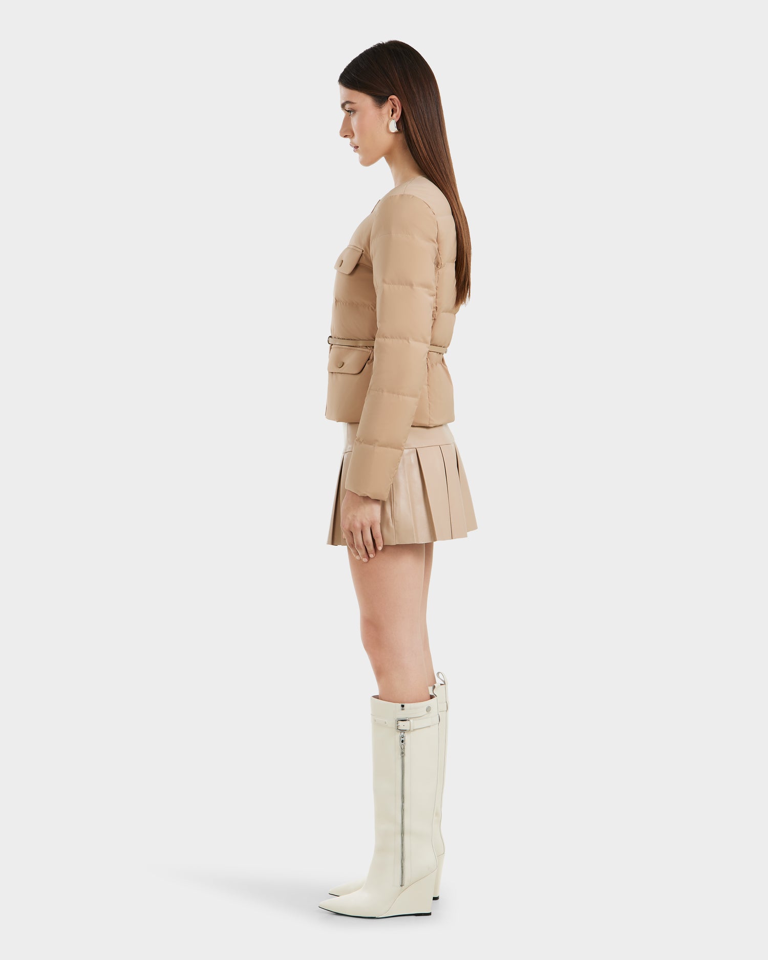 A woman stands in profile against a white background wearing Rudsak's FRANCESCA - 8125021 Almond beige quilted puffer jacket, a matching pleated mini skirt with a vegan leather belt, and knee-high white zippered boots.
