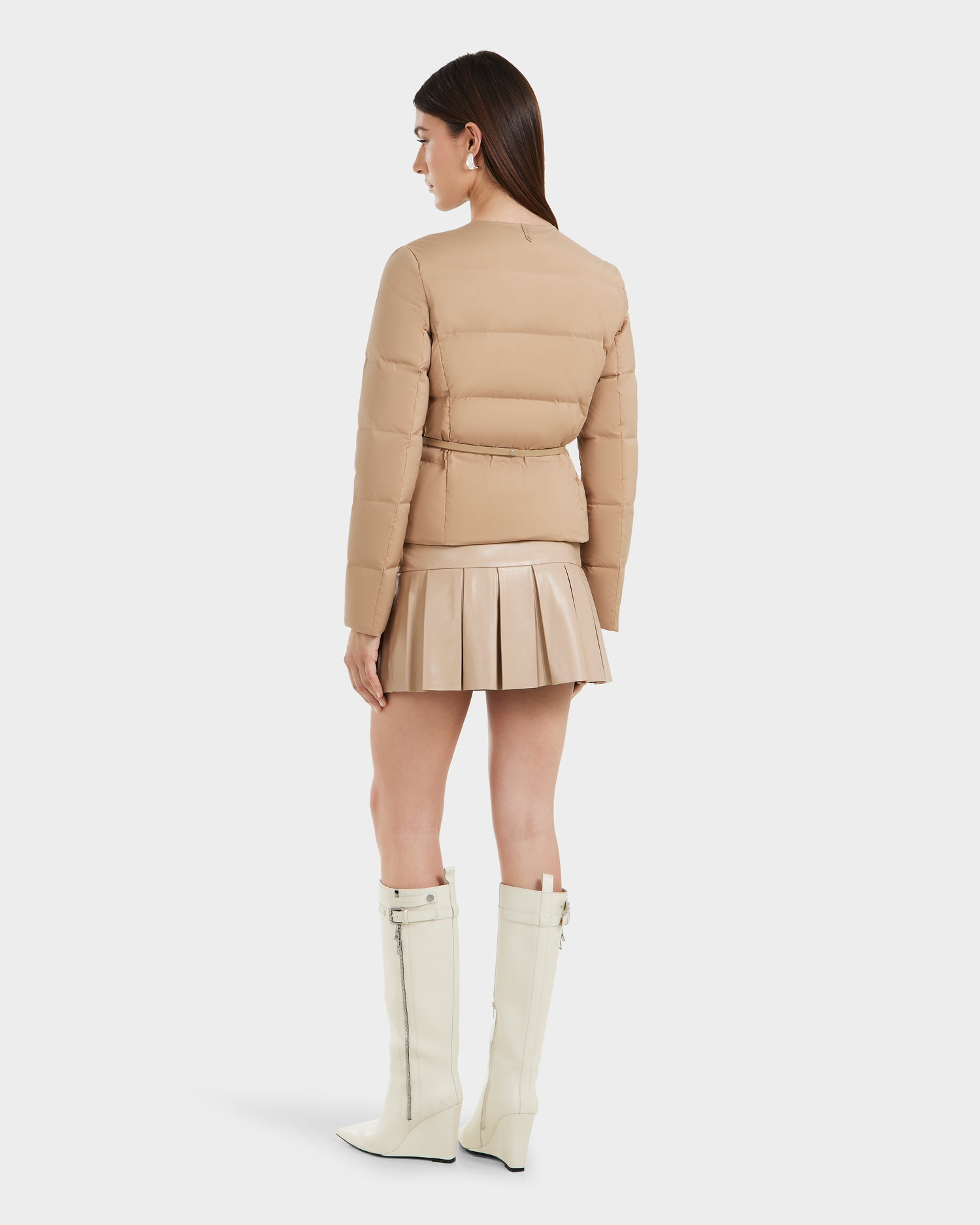 A woman faces away against a plain white background, wearing the Rudsak FRANCESCA - 8125021 Almond quilted puffer jacket, a matching pleated mini skirt, and knee-high white zippered boots.