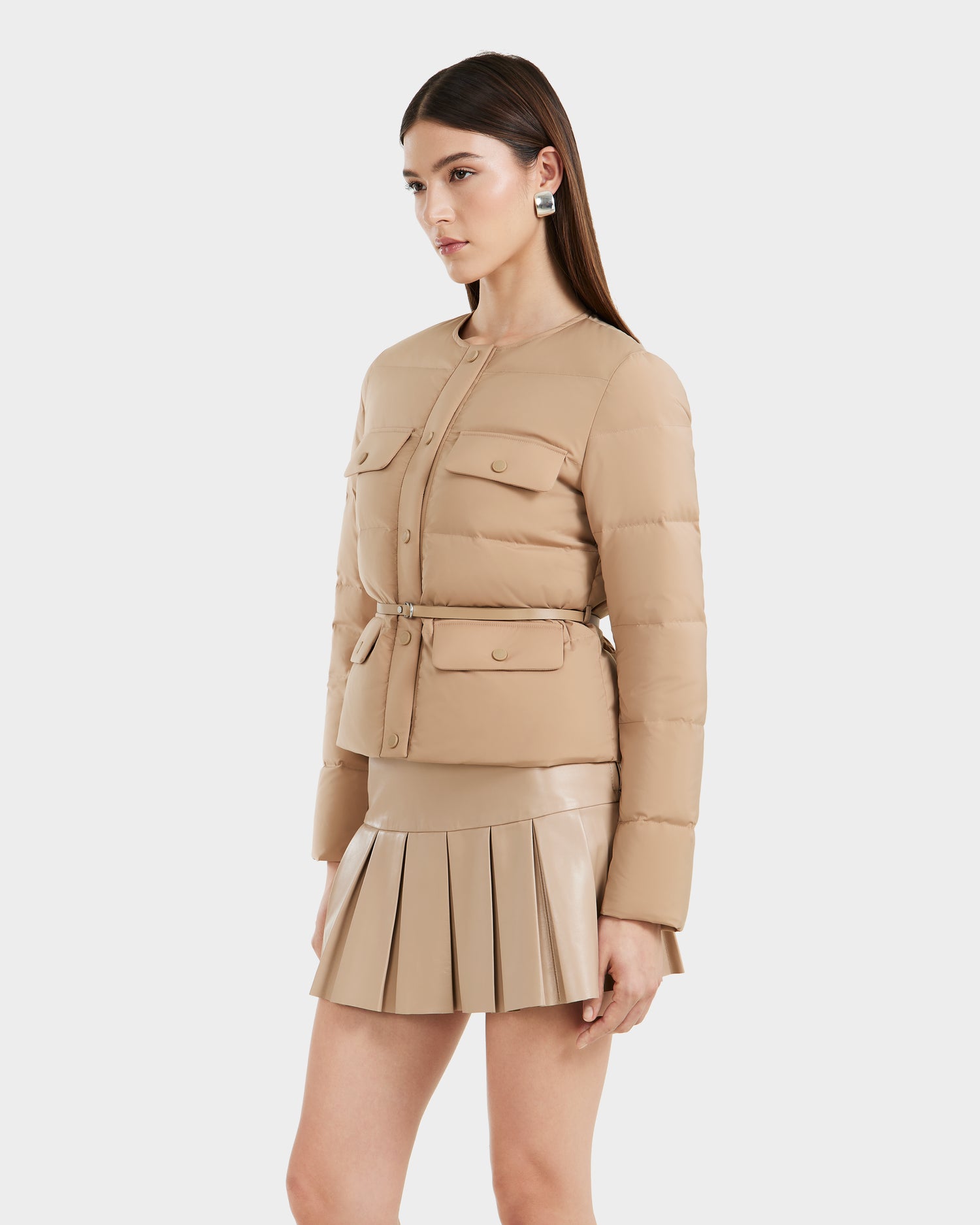A woman models the FRANCESCA - 8125021 Almond quilted puffer jacket by Rudsak with front pockets and a beige pleated mini skirt, styled with a vegan leather belt, against a plain white background.