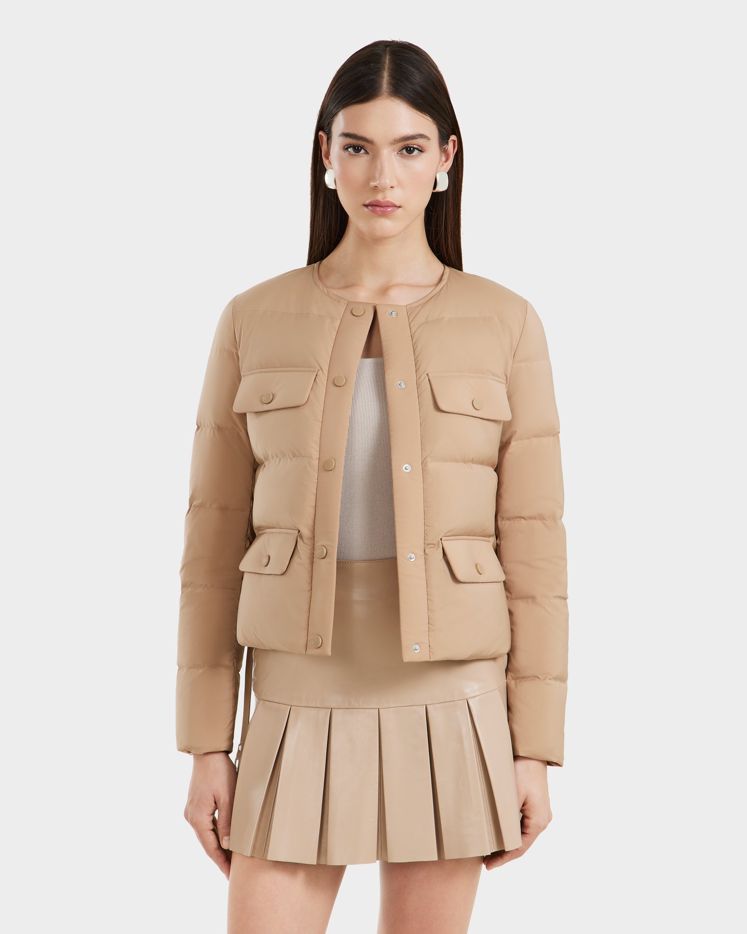 A woman with straight brown hair models the Rudsak FRANCESCA - 8125021 Almond beige quilted puffer jacket with snap buttons and flap pockets, styled over coordinating taupe and beige pieces, standing against a plain white background.