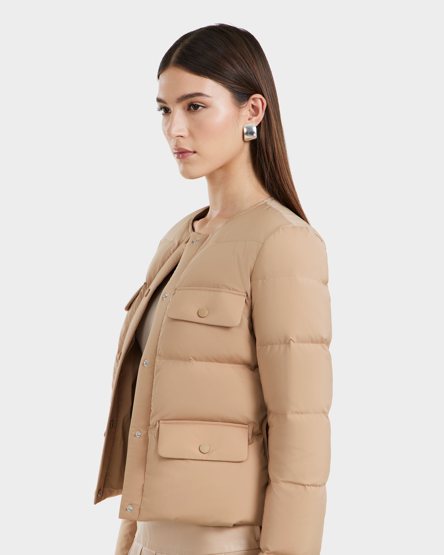A woman with straight brown hair models the Rudsak FRANCESCA - 8125021 Almond quilted puffer jacket, featuring flap pockets and silver statement earrings, posed against a white background.