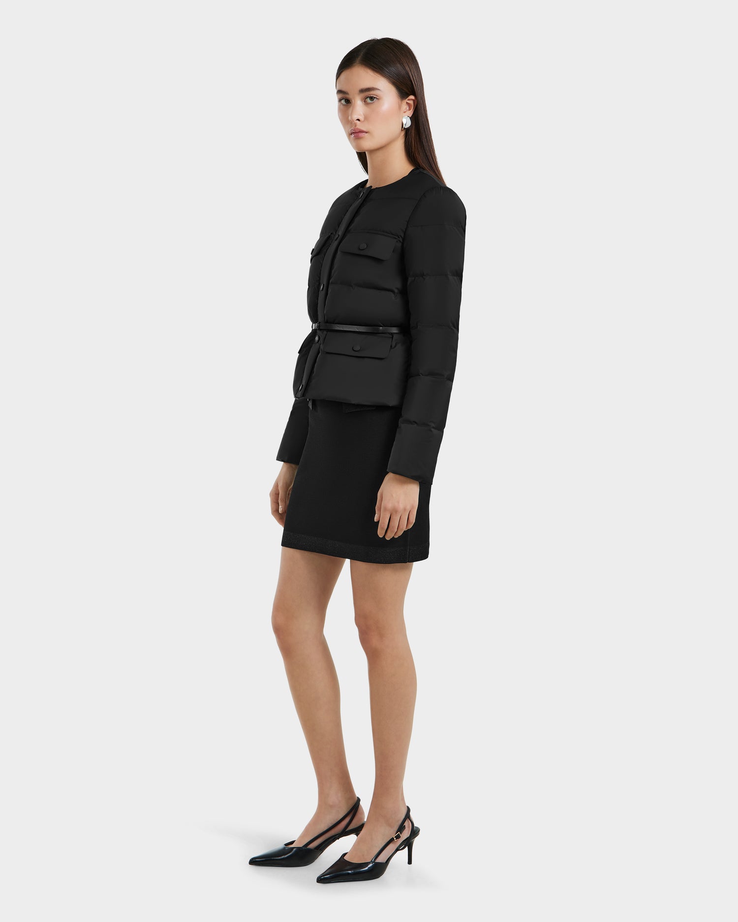 A woman with straight dark hair and a neutral expression wears the Rudsak FRANCESCA - 8125021 Black lightweight puffer jacket with a vegan leather belt, styled over a black knee-length dress, black slingback heels, and silver earrings.