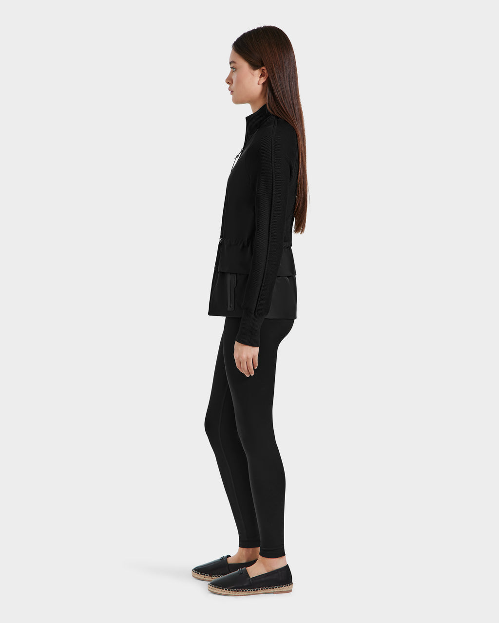 A woman with long brown hair stands in profile against a plain light gray background, wearing Rudsaks GENEVIEVE - 8125042 Black wind-resistant shell hybrid jacket—ideal for transitional weather.