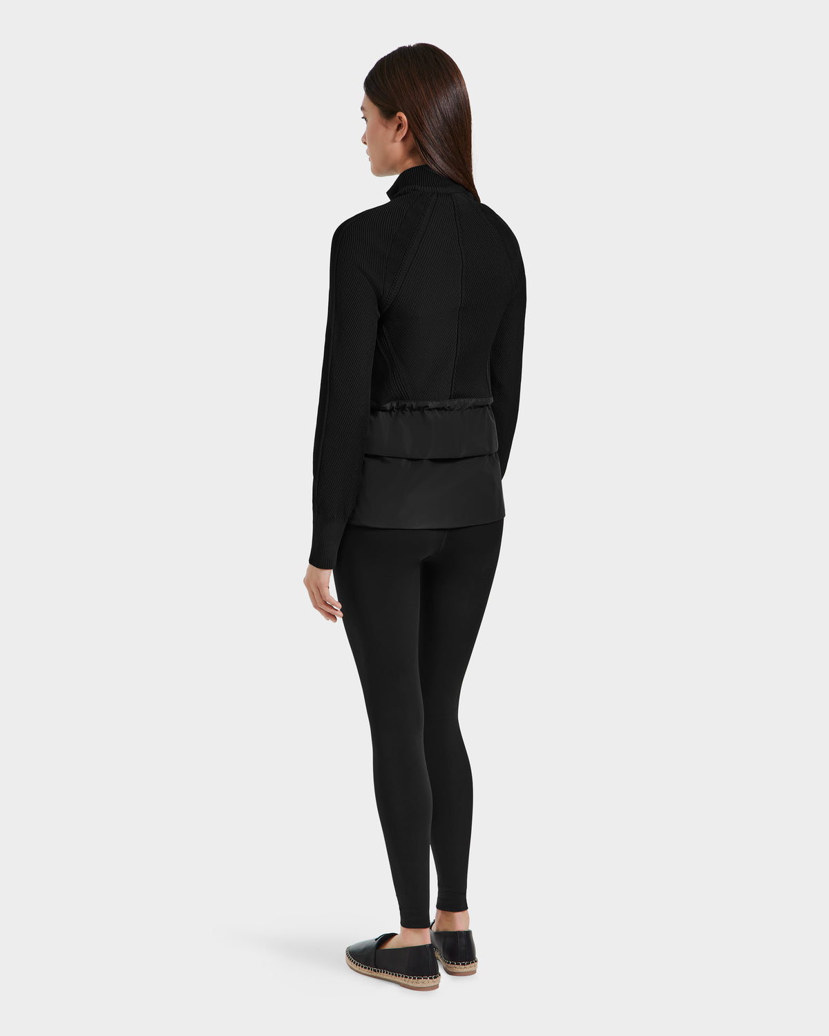 A woman with long brown hair, facing away against a white background, wears the Rudsak GENEVIEVE - 8125042 Black hybrid jacket, paired with black leggings and black shoes with beige soles.