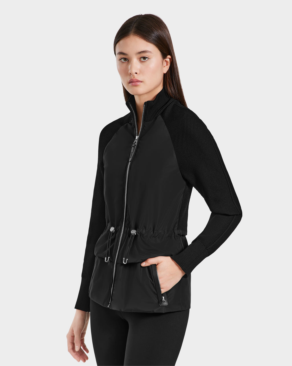 A young woman with long, straight brown hair models the Rudsak GENEVIEVE - 8125042 Black wind-resistant jacket with ribbed sleeves, front pockets, and an adjustable waist, paired with black leggings against a light gray background.