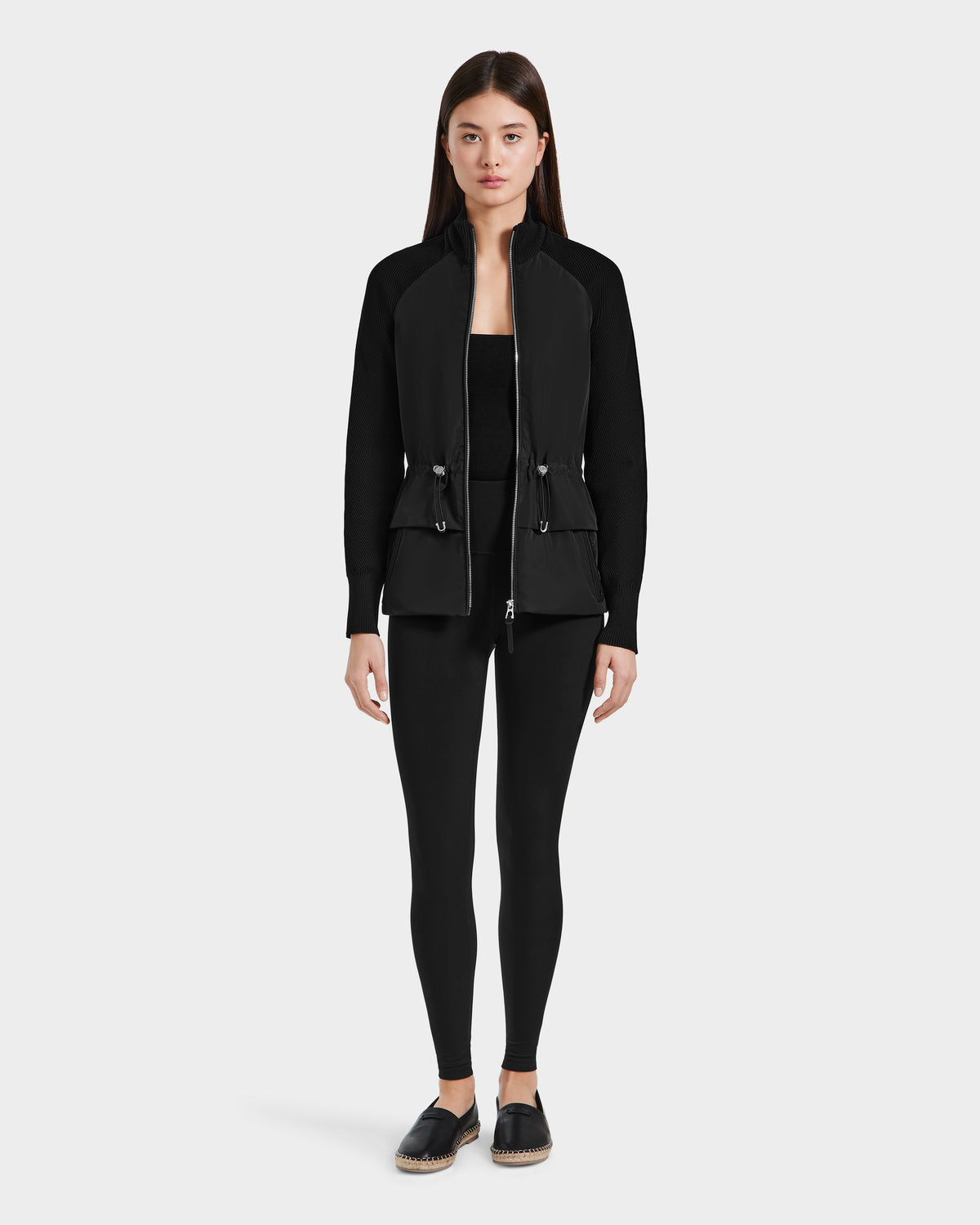 A woman with straight brown hair and a neutral expression stands facing forward in the Rudsak GENEVIEVE - 8125042 Black hybrid jacket, paired with black leggings and slip-on shoes, posed against a plain white background.
