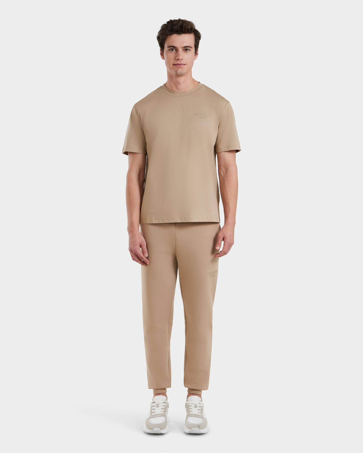 A person stands facing forward in Rudsak’s GRANT - 6525036 Almond, a matching light brown men’s crewneck tee and jogger pants set, paired with white sneakers against a plain white background.