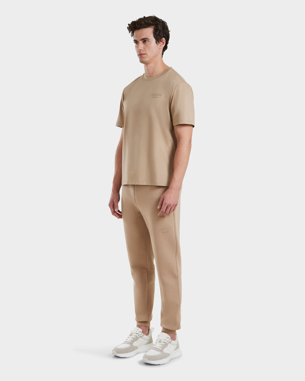 A man stands against a plain white background wearing the Rudsak GRANT - 6525036 Almond premium cotton crewneck tee and matching jogger set, paired with white and beige sneakers.