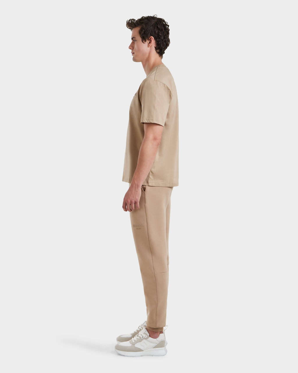 A man in profile faces left, wearing the GRANT - 6525036 Almond crewneck tee and matching jogger pants by Rudsak, paired with white sneakers, all set against a plain white background.