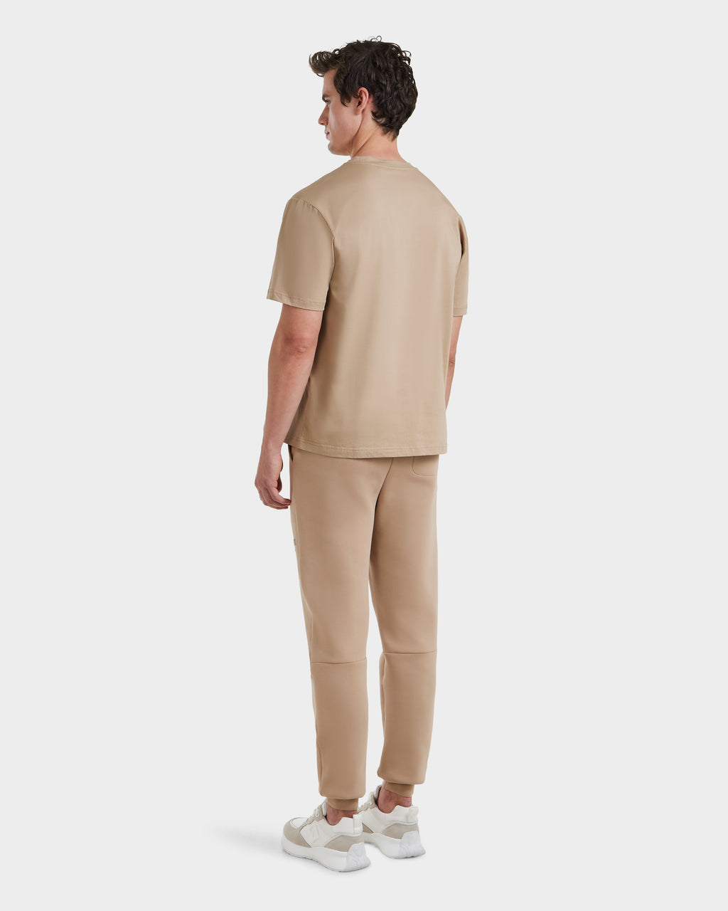 A man stands facing away, dressed in the Rudsak GRANT - 6525036 Almond premium cotton crewneck tee, matching beige pants, and white sneakers, set against a plain white background.