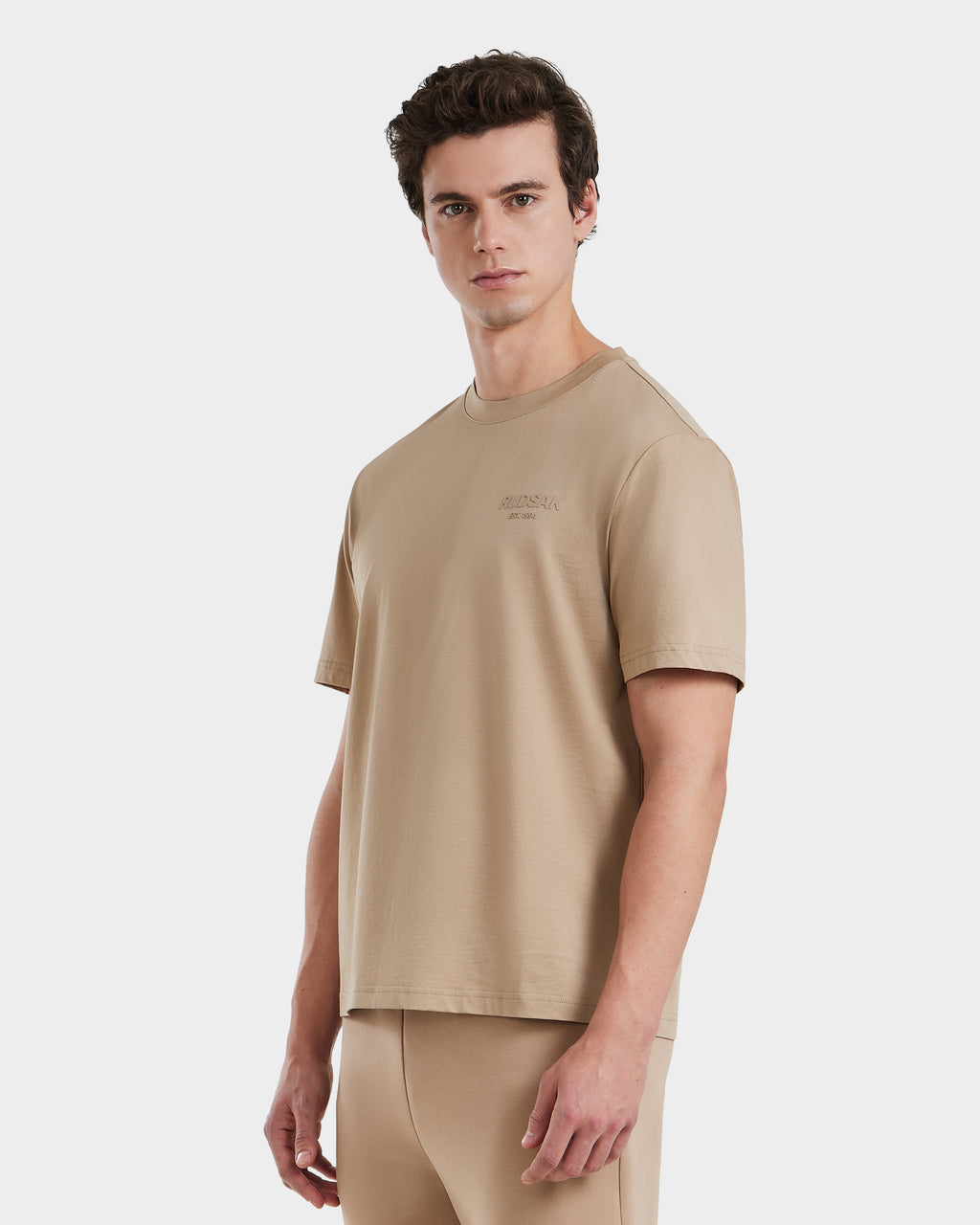 A young man models the Rudsak GRANT - 6525036 Almond crewneck tee and matching pants against a plain white background, looking directly at the camera with a neutral expression.