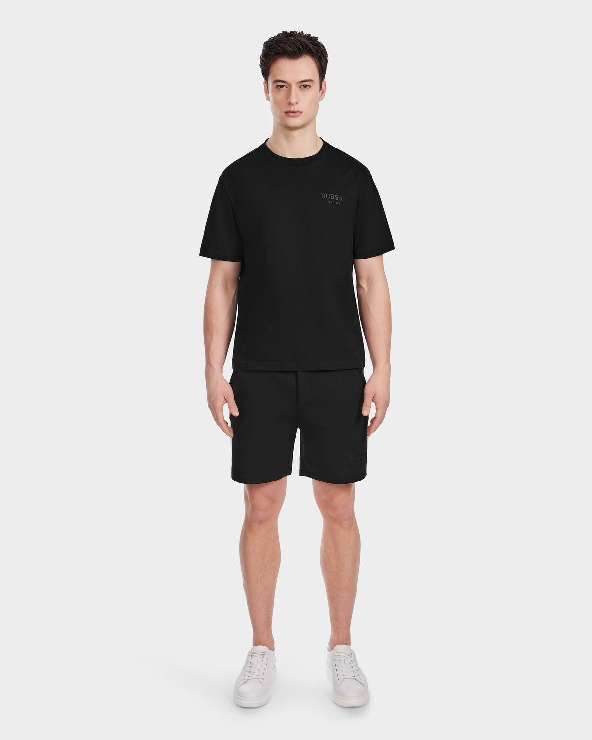 A person stands facing forward, wearing the Rudsak GRANT - 6525036 Black men’s tee, black shorts, and white sneakers against a plain white background.