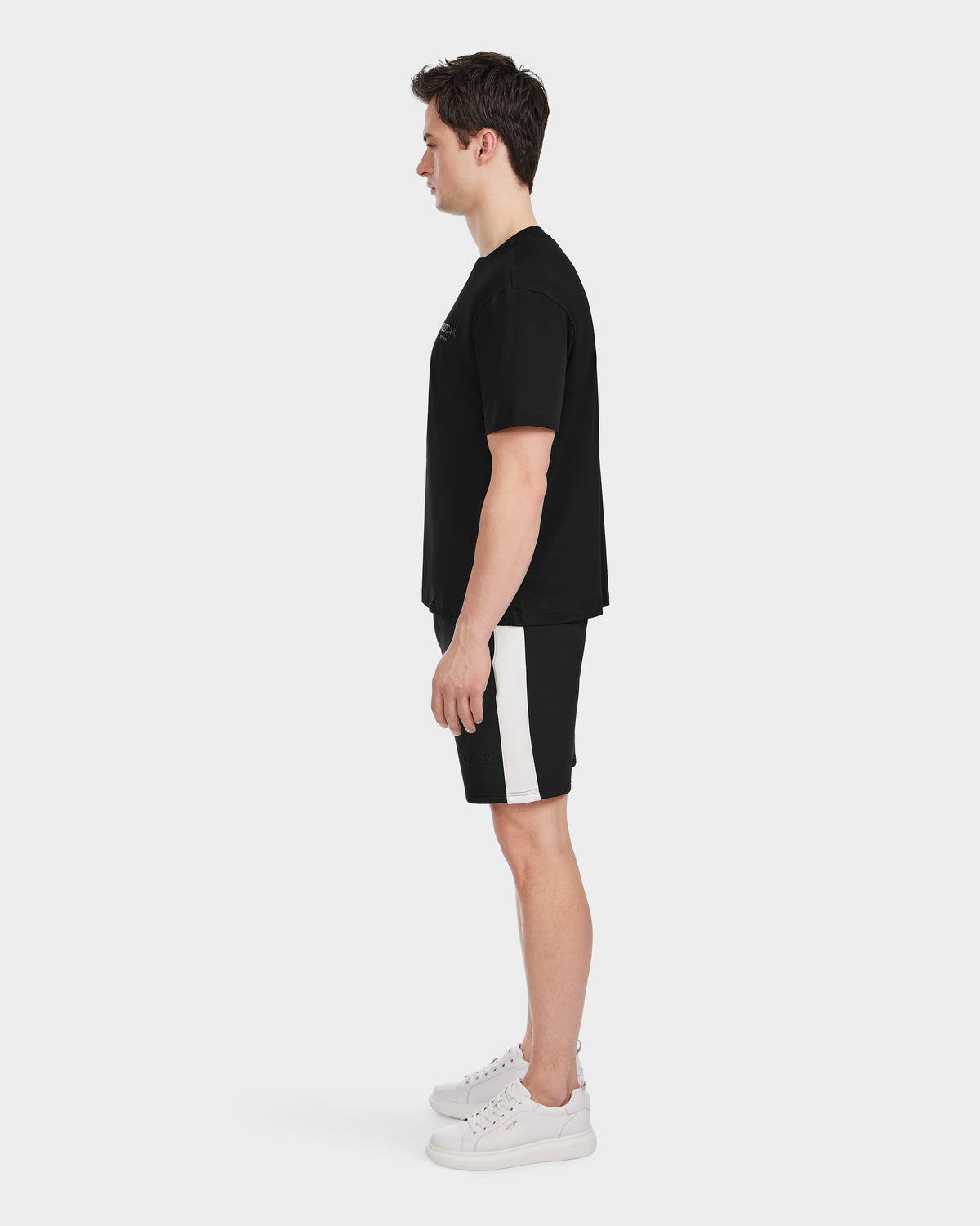 A man stands in profile facing left, wearing the Rudsak GRANT - 6525036 Black crewneck men’s tee, black shorts with a white side stripe, and white sneakers against a plain white background.