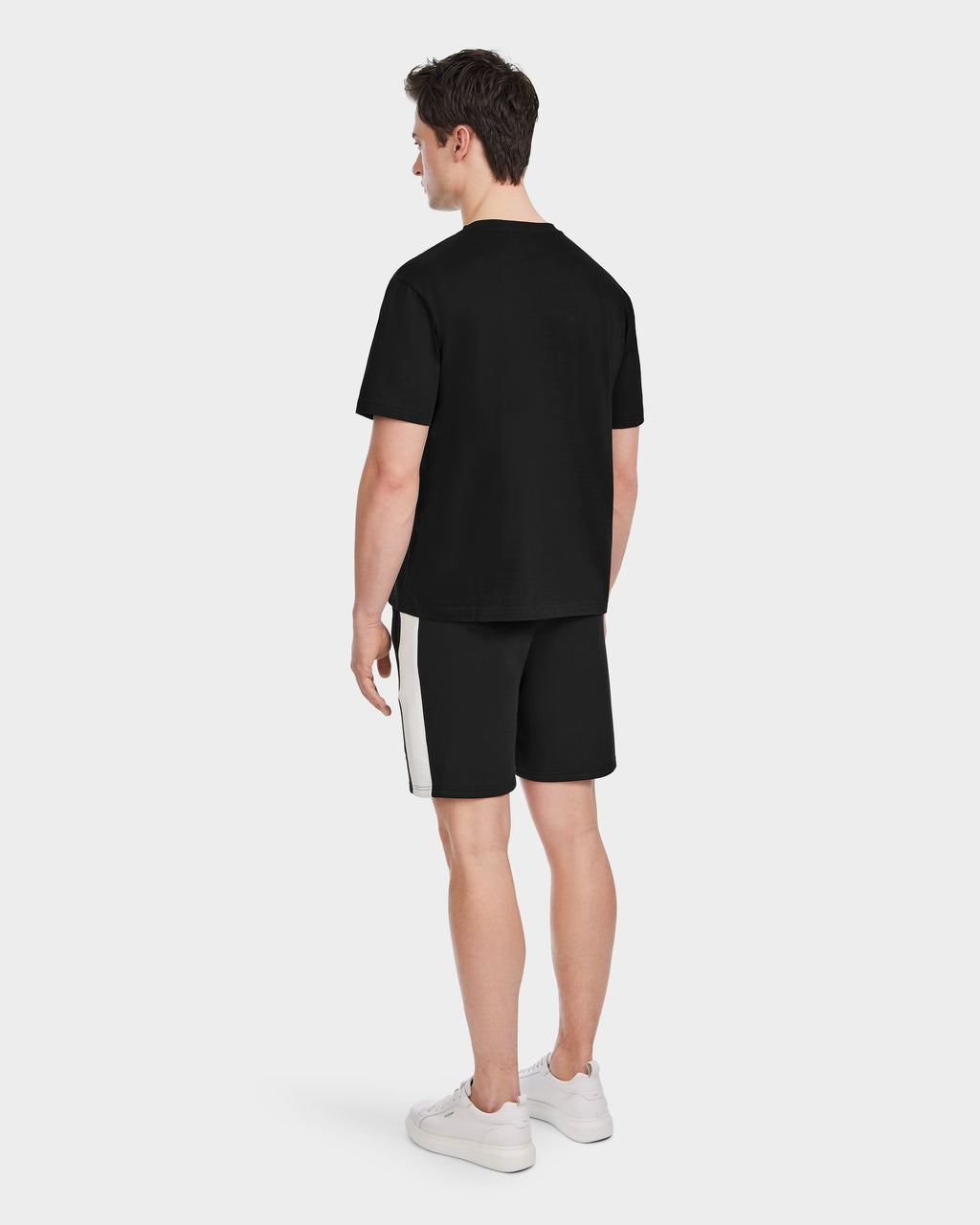 A man, seen from behind, wears the Rudsak GRANT - 6525036 Black premium cotton crewneck men’s tee with black shorts featuring a white side stripe and white sneakers against a plain light background.