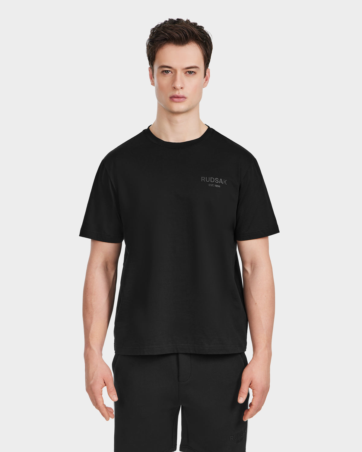A young man faces forward, wearing the Rudsak GRANT - 6525036 Black men’s tee, made from premium cotton with a classic crewneck and RUDSAK subtly printed on the left chest. The background is white.