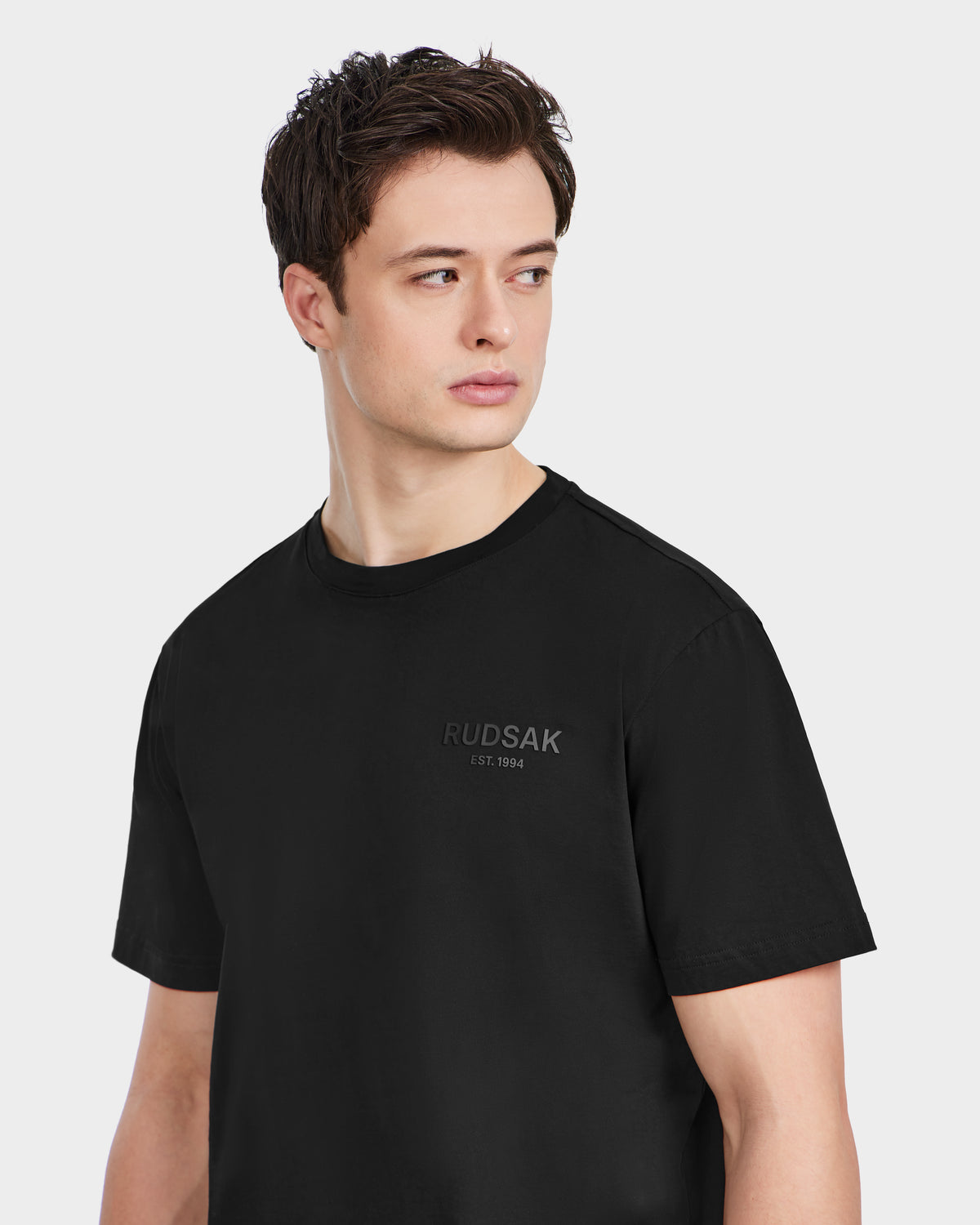 A young man with short dark hair wears the Rudsak GRANT - 6525036 Black premium cotton crewneck tee, featuring RUDSAK EST. 1994 on the chest, as he looks to his left against a plain light background.