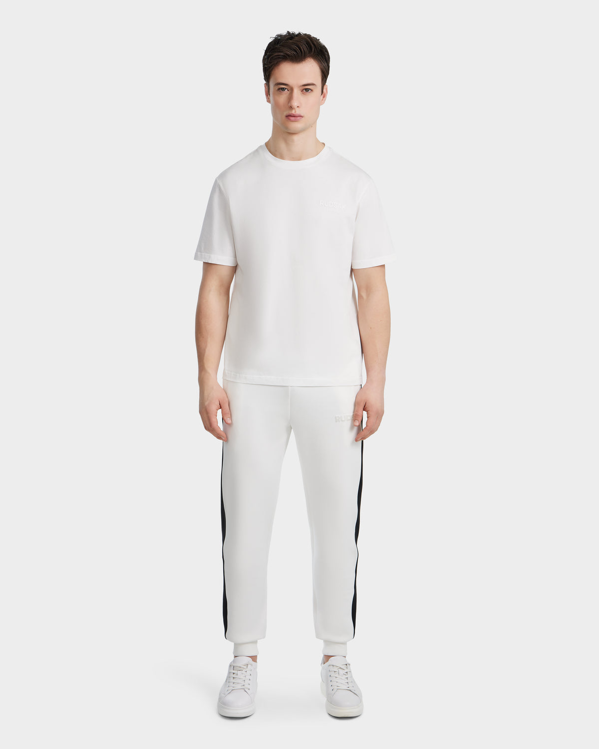 A man faces forward, wearing the Rudsak GRANT - 6525036 Iceland white men’s crewneck tee and matching jogger pants with black side stripes, paired with white sneakers against a plain light background.