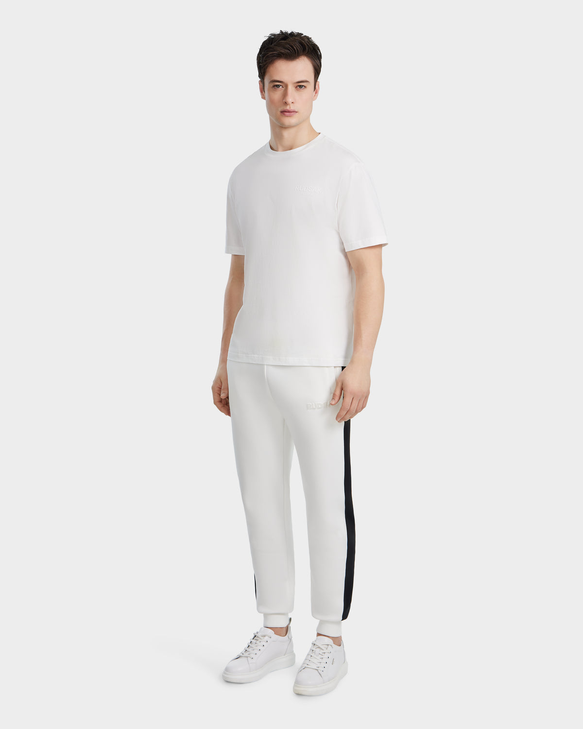 A man faces forward against a light background, wearing the Rudsak GRANT - 6525036 Iceland premium cotton mens crewneck tee, white jogger-style pants with black side stripes, and white sneakers.
