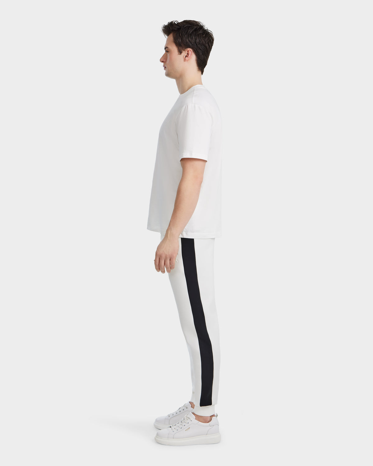 A man in profile faces left, dressed in the Rudsak GRANT - 6525036 Iceland premium white cotton crewneck tee, paired with white jogger pants featuring a black side stripe and white sneakers, set against a plain white background.