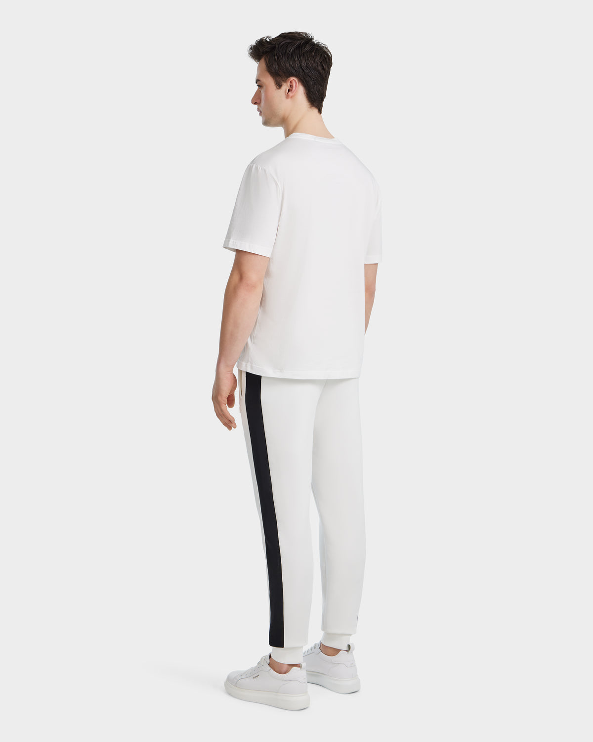 A man faces away, wearing the Rudsak GRANT - 6525036 Iceland mens premium cotton crewneck tee with white pants featuring a black side stripe and white sneakers, set against a plain white background.