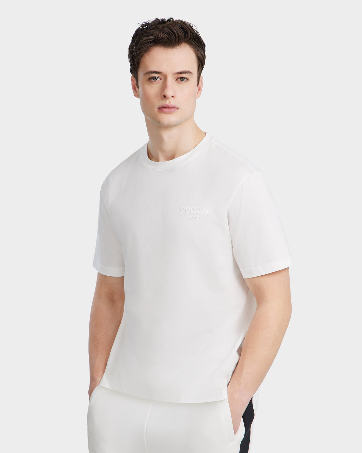 A man with short dark hair models the Rudsak GRANT - 6525036 Iceland mens white crewneck tee and matching pants, standing hands-in-pockets against a white background, looking directly at the camera.