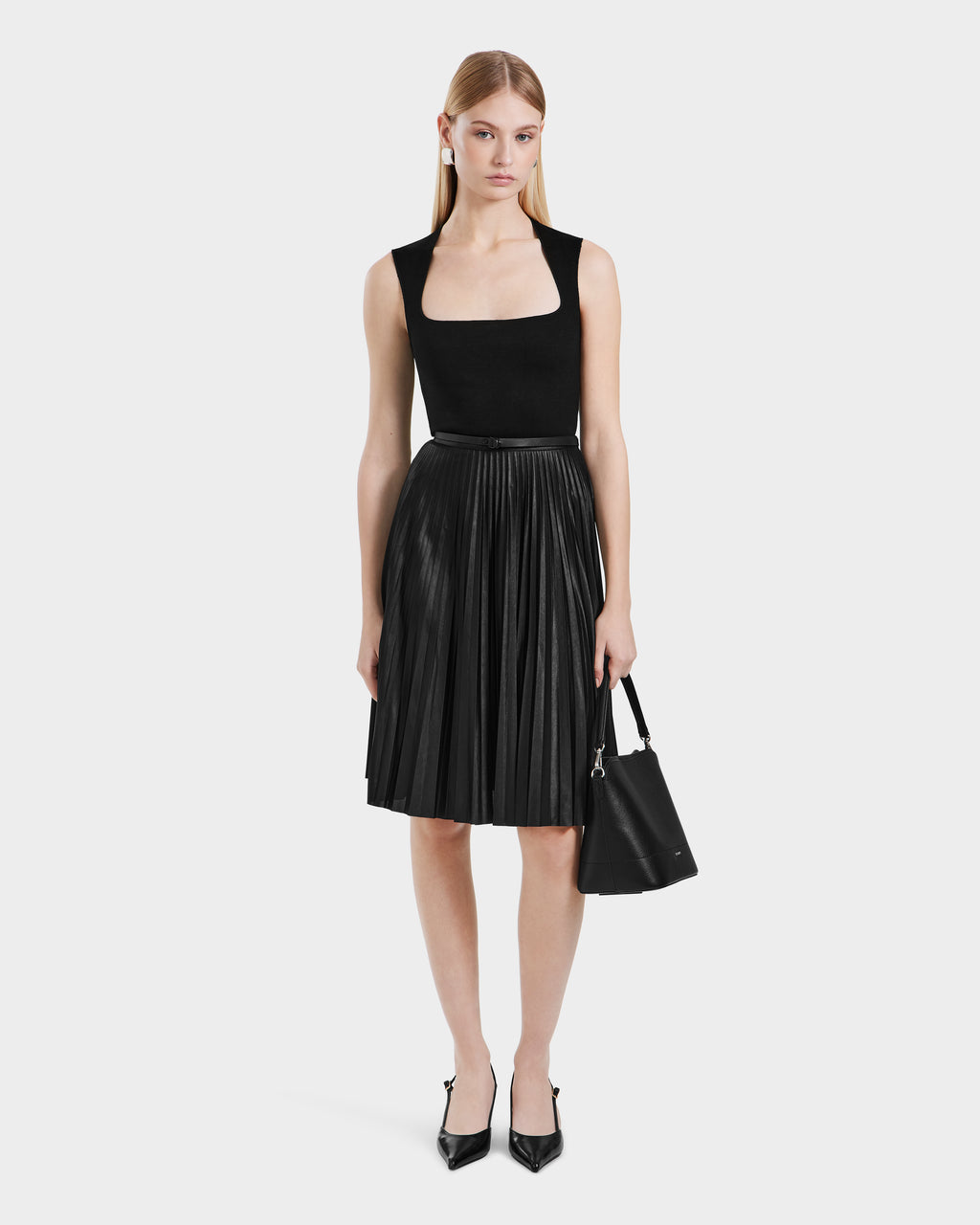 A woman faces forward in a black sleeveless top and the JANE - 8525029 Black pleated midi skirt by Rudsak, styled with black pointed-toe heels and a black handbag, set against a plain white background.