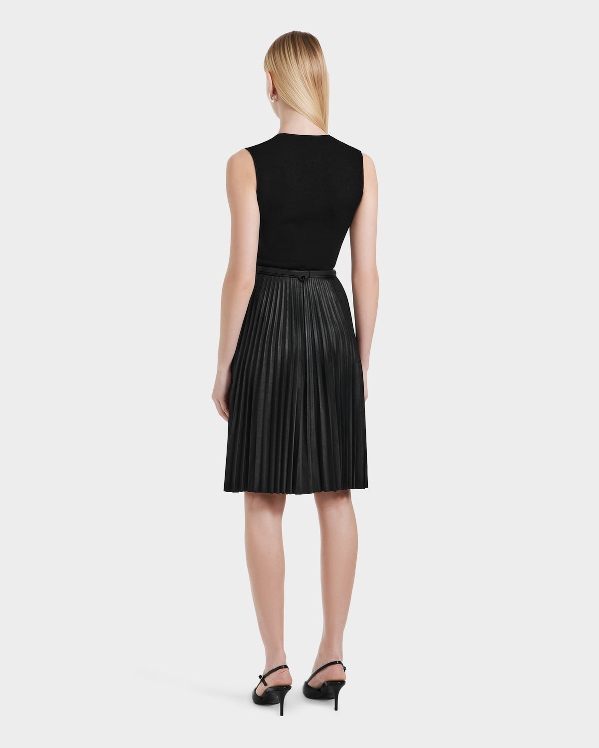 A woman with straight blonde hair styled down stands facing away in a sleeveless black top, black heeled sandals, and the Rudsak JANE - 8525029 Black pleated midi skirt, set against a plain white background.