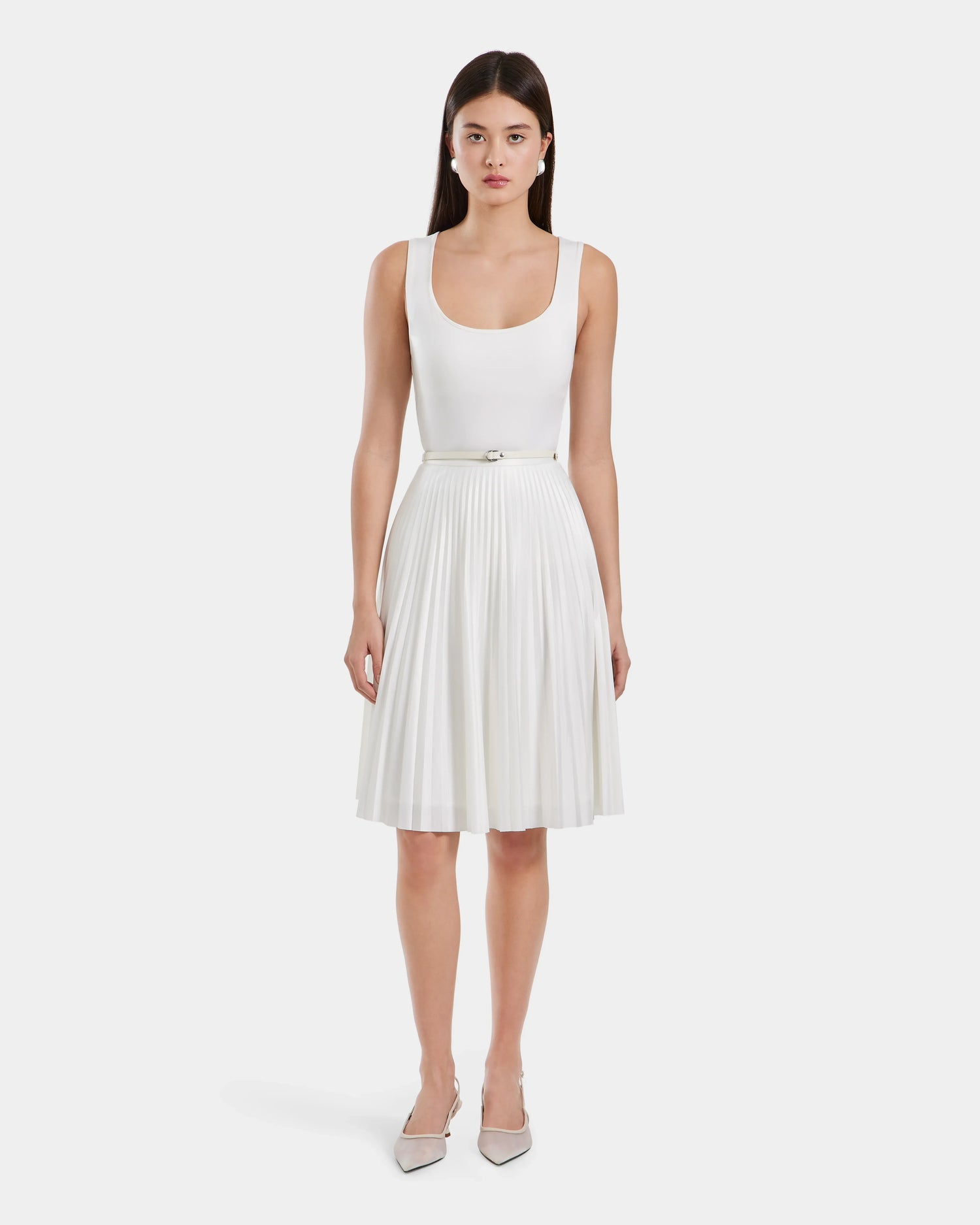 A woman with straight dark hair and a neutral expression faces forward, wearing the JANE - 8525029 Iceland sleeveless white knee-length dress by Rudsak with a pleated midi skirt and thin belt, styled with pointed-toe heels against a plain white background.