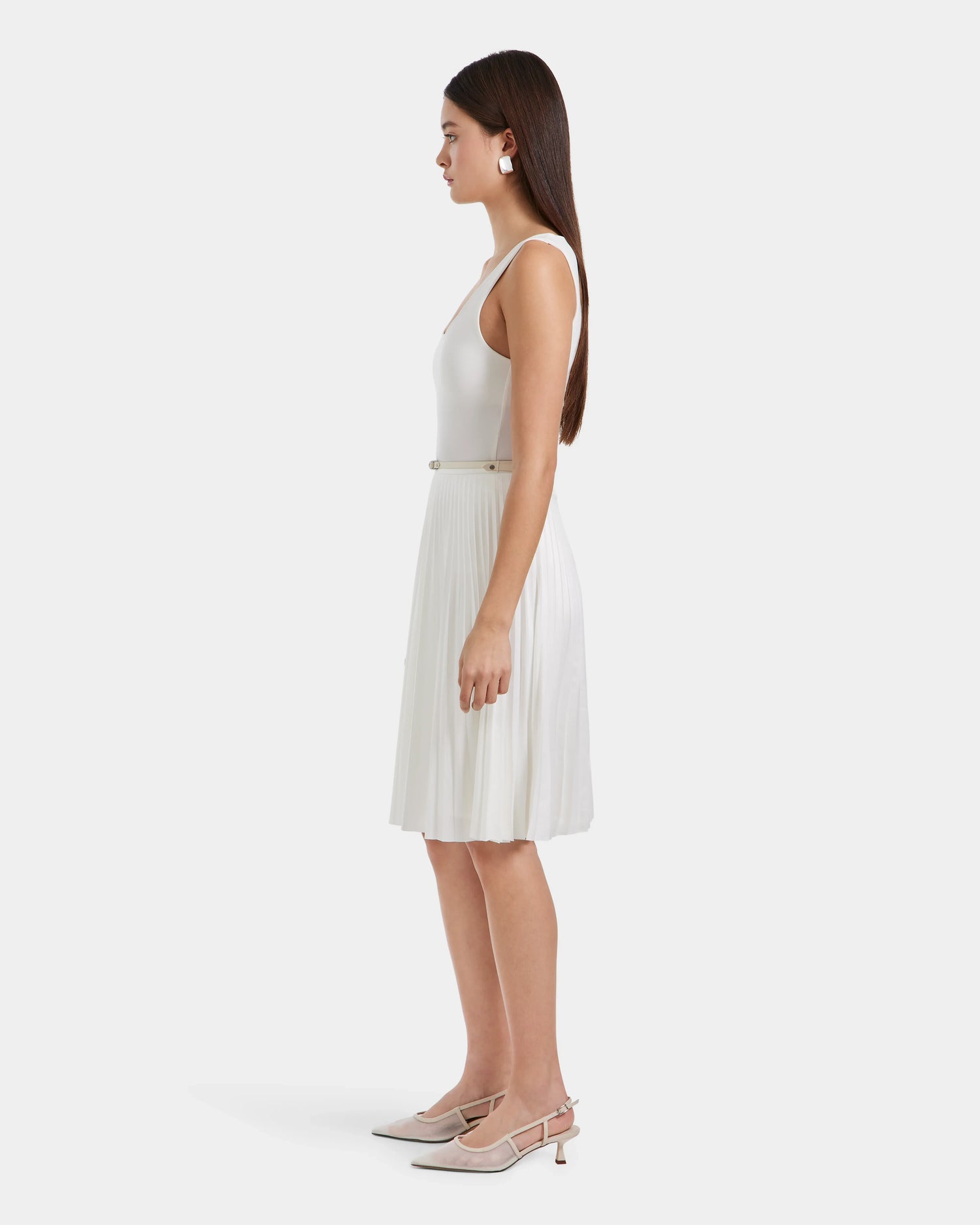 A woman with long straight hair stands in profile against a plain background, wearing the JANE - 8525029 Iceland sleeveless white top by Rudsak, a women’s pleated midi skirt, light-colored heels, and white earrings.