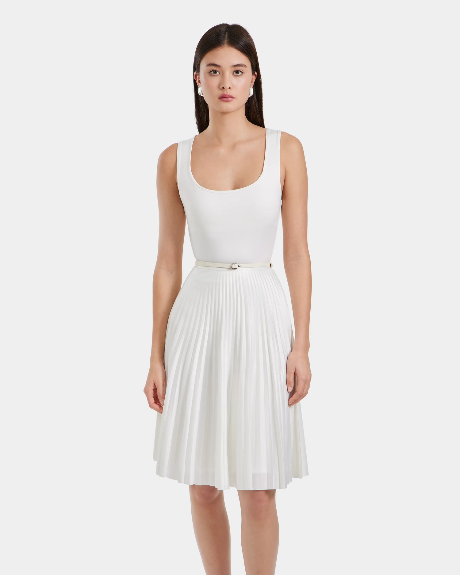 A woman with long, dark hair wears the Rudsak JANE - 8525029 Iceland sleeveless white top and pleated skirt with belt, standing against a plain light background.