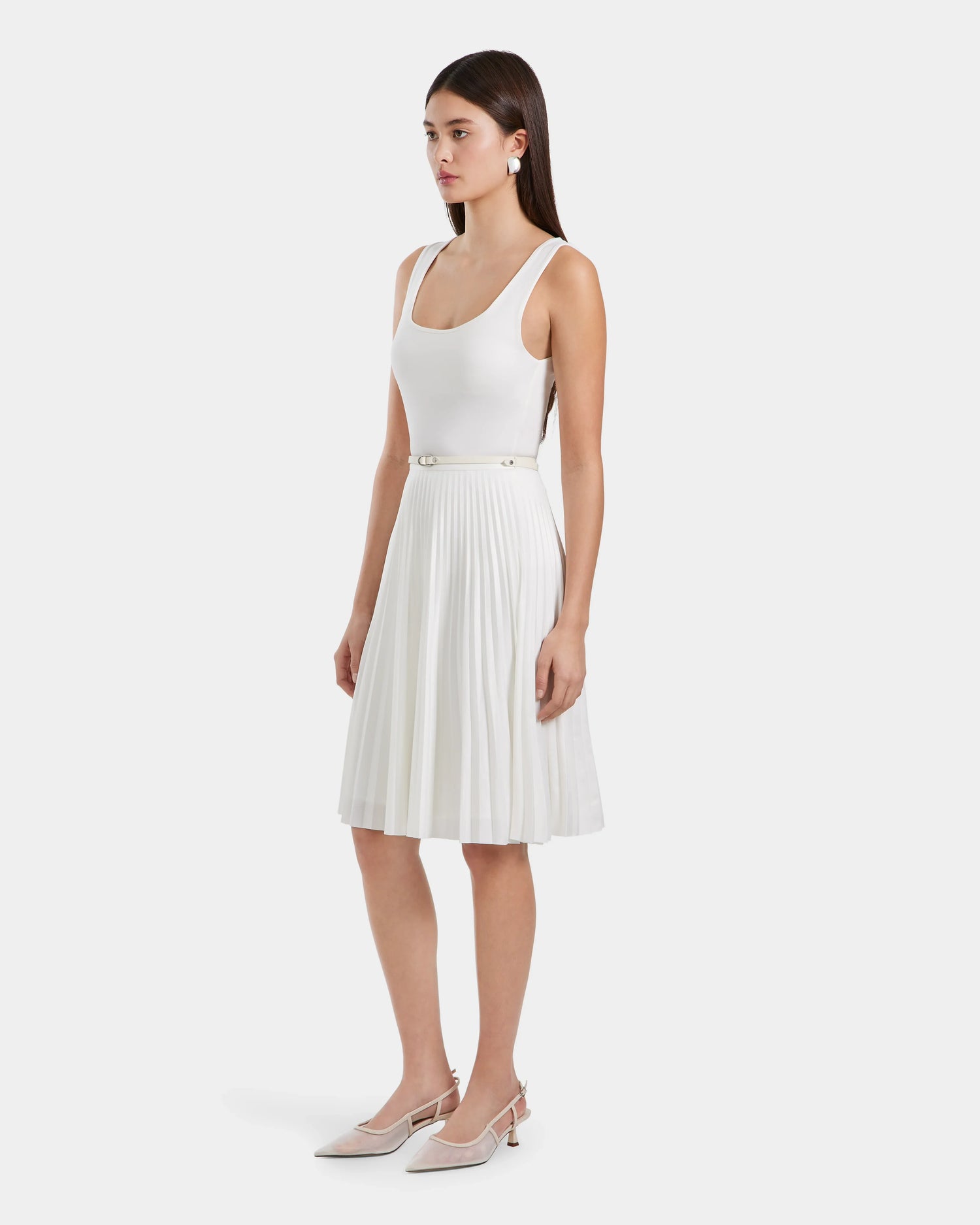 A woman in profile wears the Rudsak JANE - 8525029 Iceland sleeveless white scoop-neck top, a pleated midi skirt with a slim belt, and white pointed-toe slingback heels. She has straight brown hair and minimal accessories.