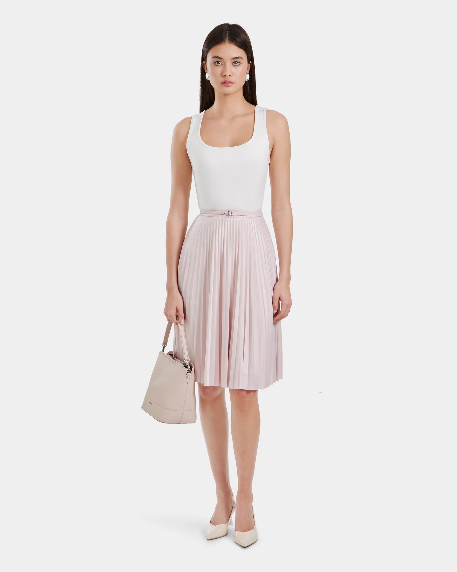 A woman with straight brown hair and a neutral expression stands facing forward, wearing Rudsak’s JANE - 8525029 Light Pink women’s pleated midi skirt, a sleeveless white top, cream heels, and holding a beige handbag.