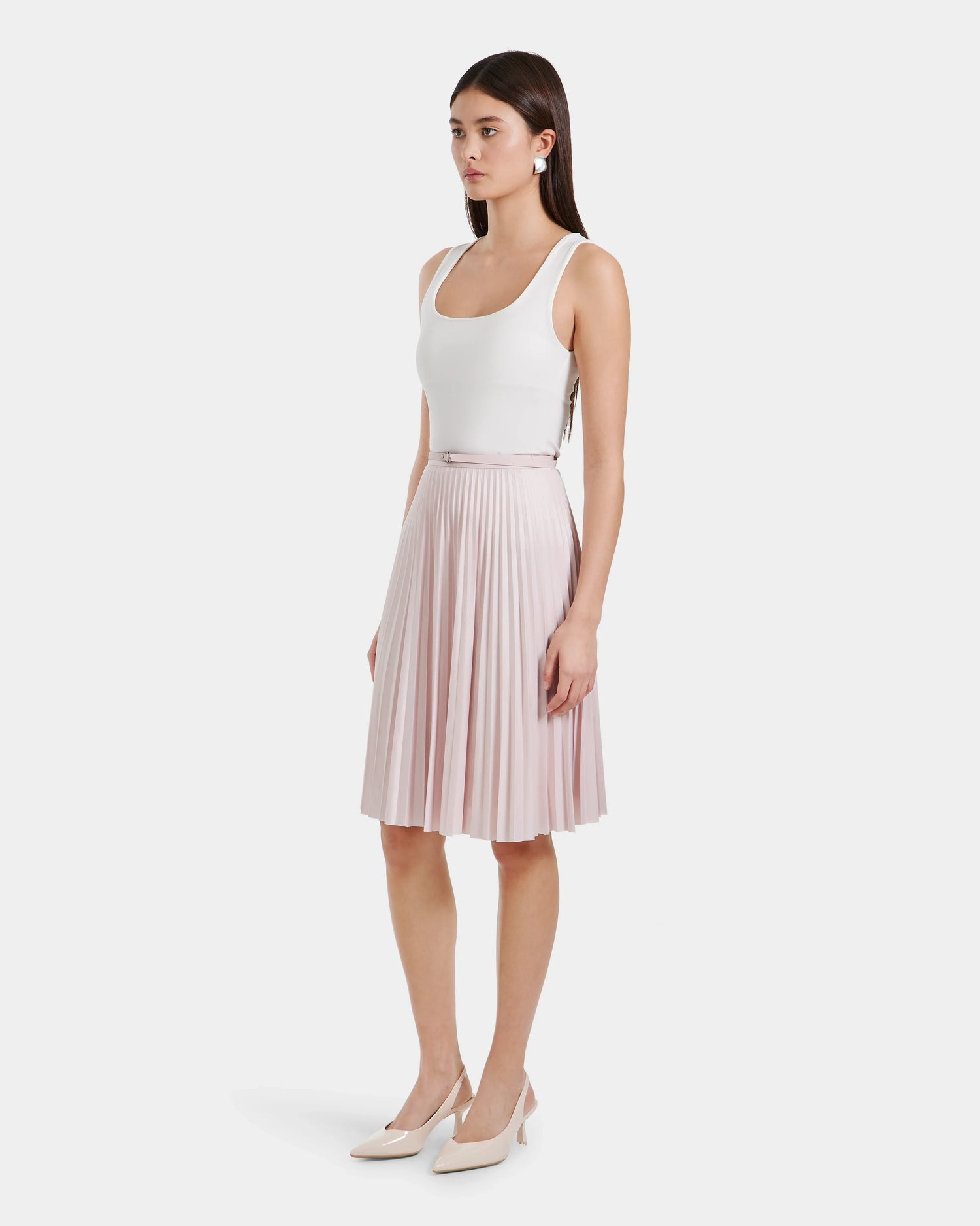 A woman stands in profile against a plain white background, wearing a white sleeveless top, nude heels, and the Rudsak JANE - 8525029 midi skirt in light pink.