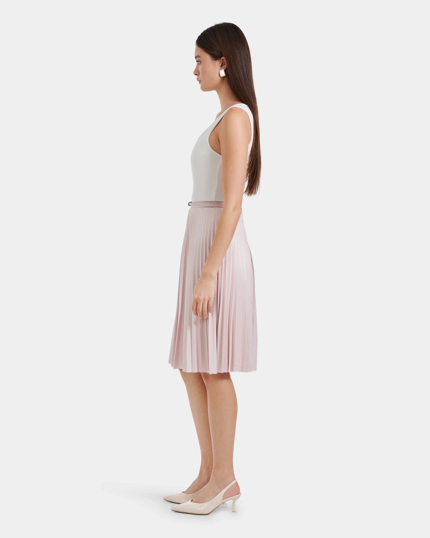 A woman stands in profile against a white background, wearing a sleeveless white top, chic Rudsak JANE - 8525029 Light Pink midi skirt, white earrings, and pale pink slingback heels. Her straight hair falls past her shoulders.