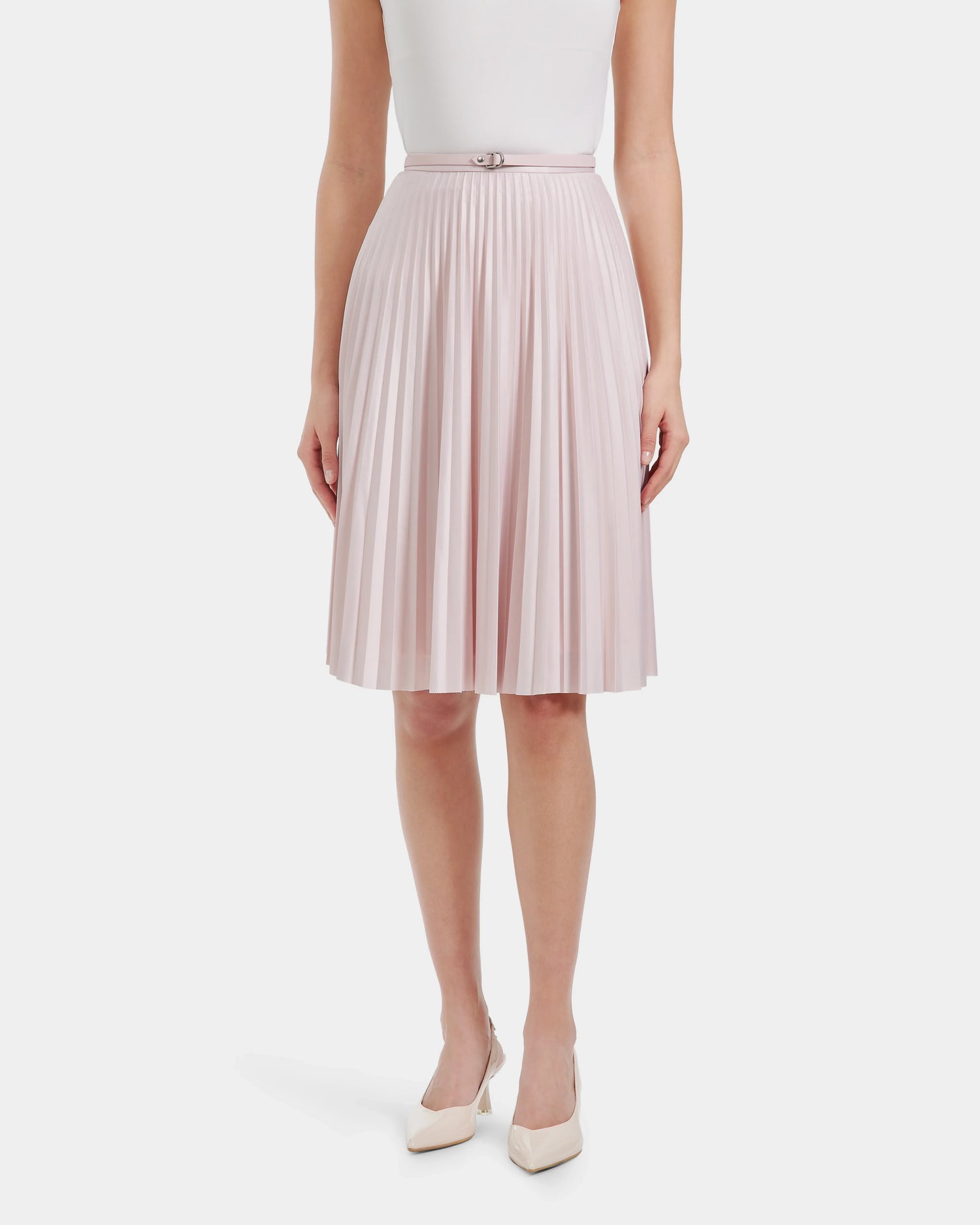 A person wears Rudsaks JANE - 8525029 Light Pink flowy pleated skirt with a thin belt, paired with a white sleeveless top and light-colored pointed-toe heels, standing against a plain white background.
