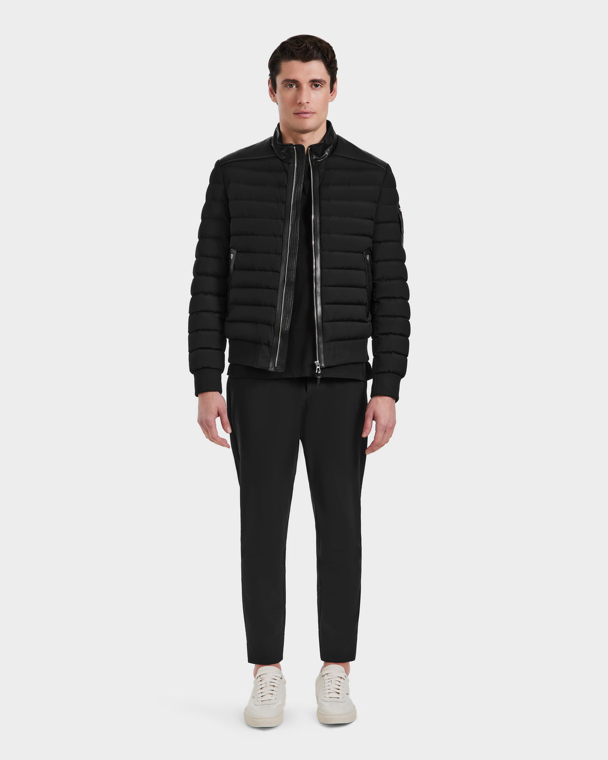 A man faces forward wearing the Rudsak JOSHUA - 6124542 Black mens biker down jacket, paired with black pants and white sneakers, set against a plain white background.